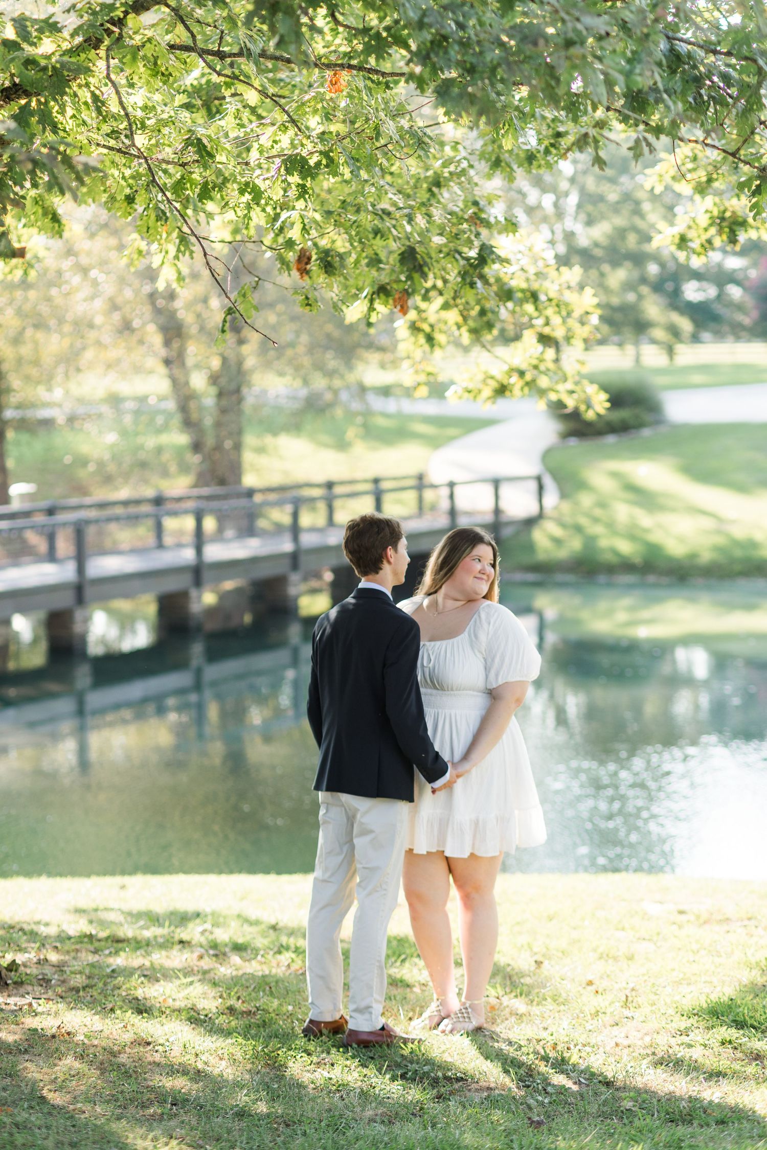 Couple holds hands by a sunlit pond with a wooden bridge, man in navy blazer, woman in white mini dress.