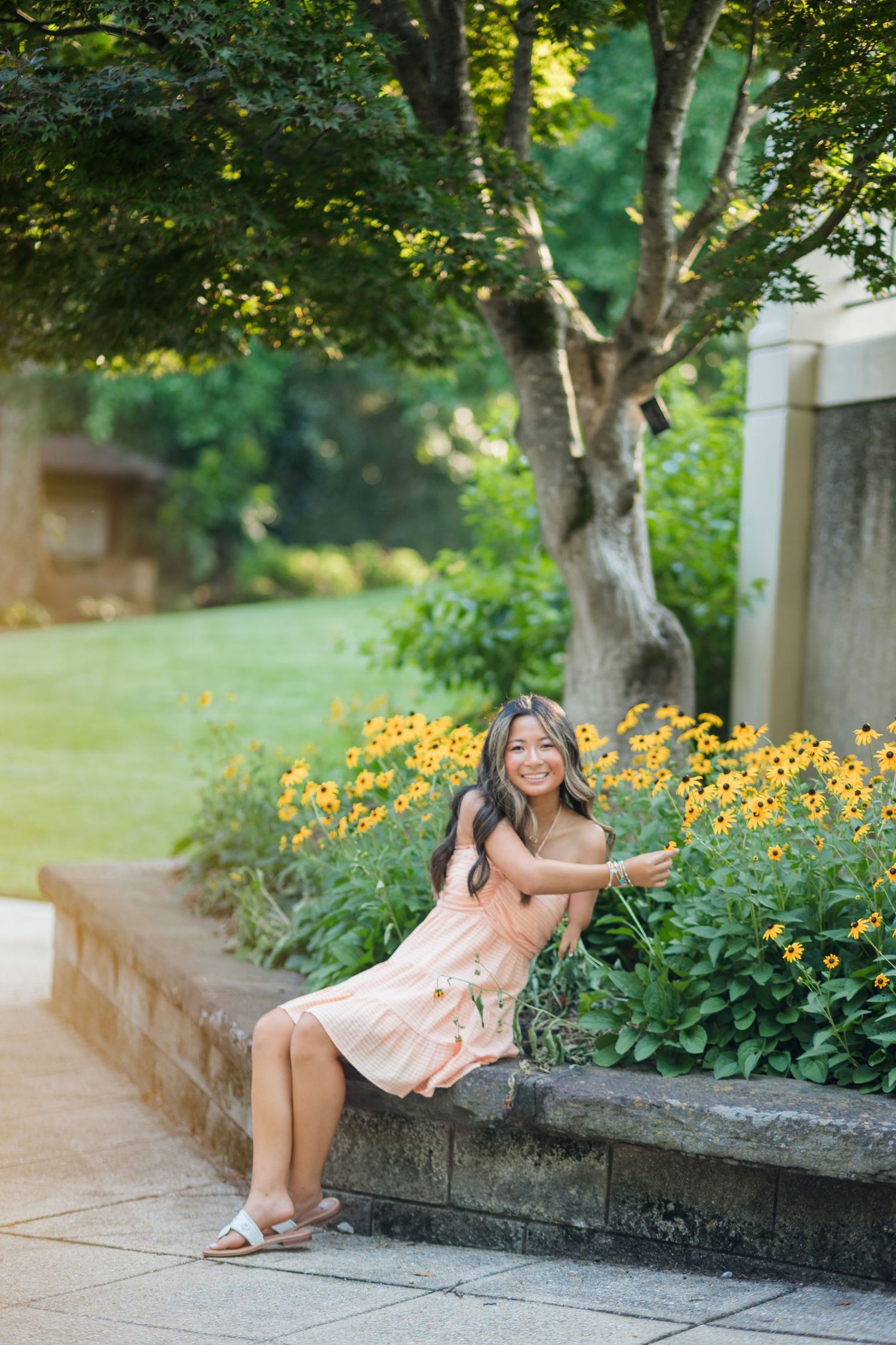 Young woman in peach dress sitting on stone garden wall surrounded by yellow flowers.