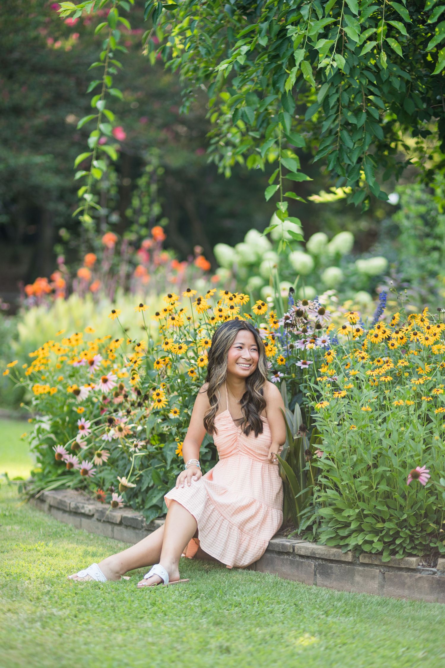 Young woman in peach dress sitting by colorful garden flowers, smiling outdoors.