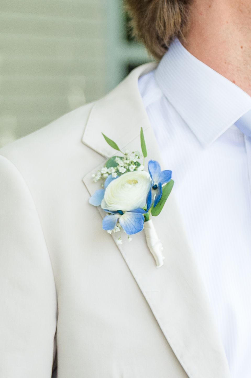 Close-up of a groom's white suit lapel adorned with a boutonniere featuring white ranunculus and blue hydrangea blooms.