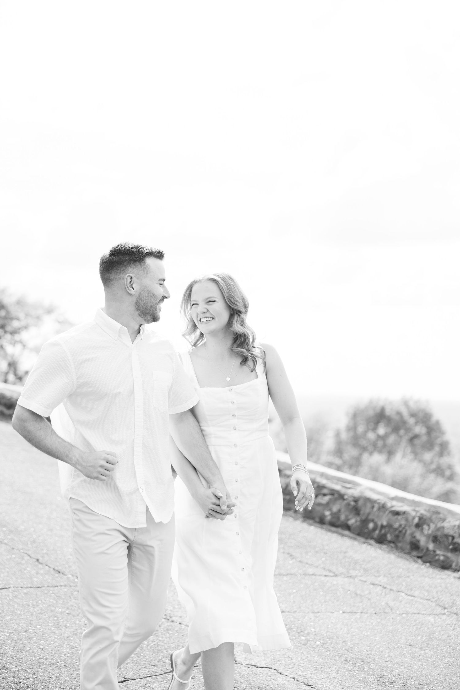 Black and white photo of a smiling couple holding hands and walking together outdoors on a sunny day.