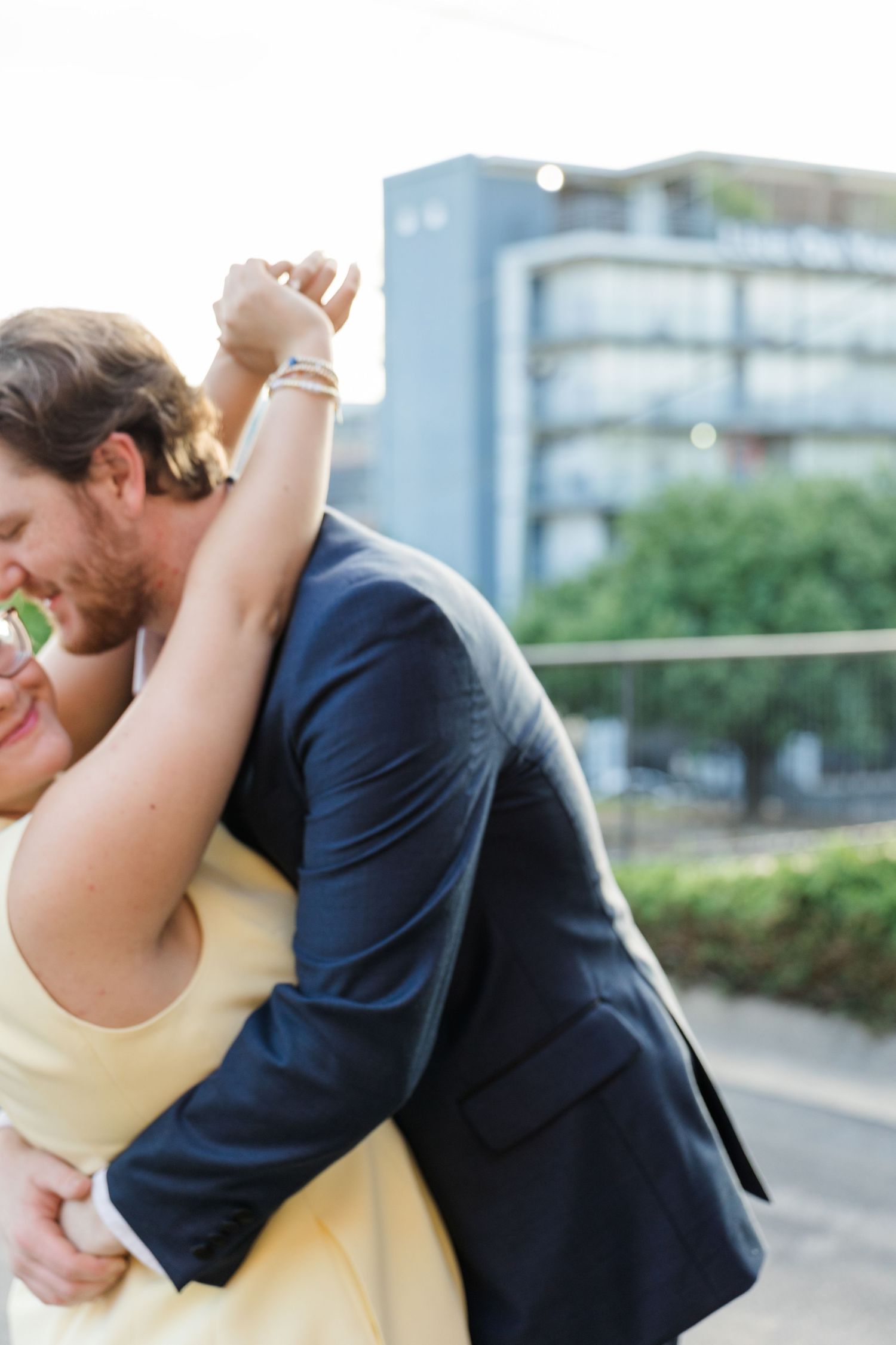 Couple embracing outdoors, woman in yellow dress and man in navy suit, sharing a romantic moment with urban backdrop.