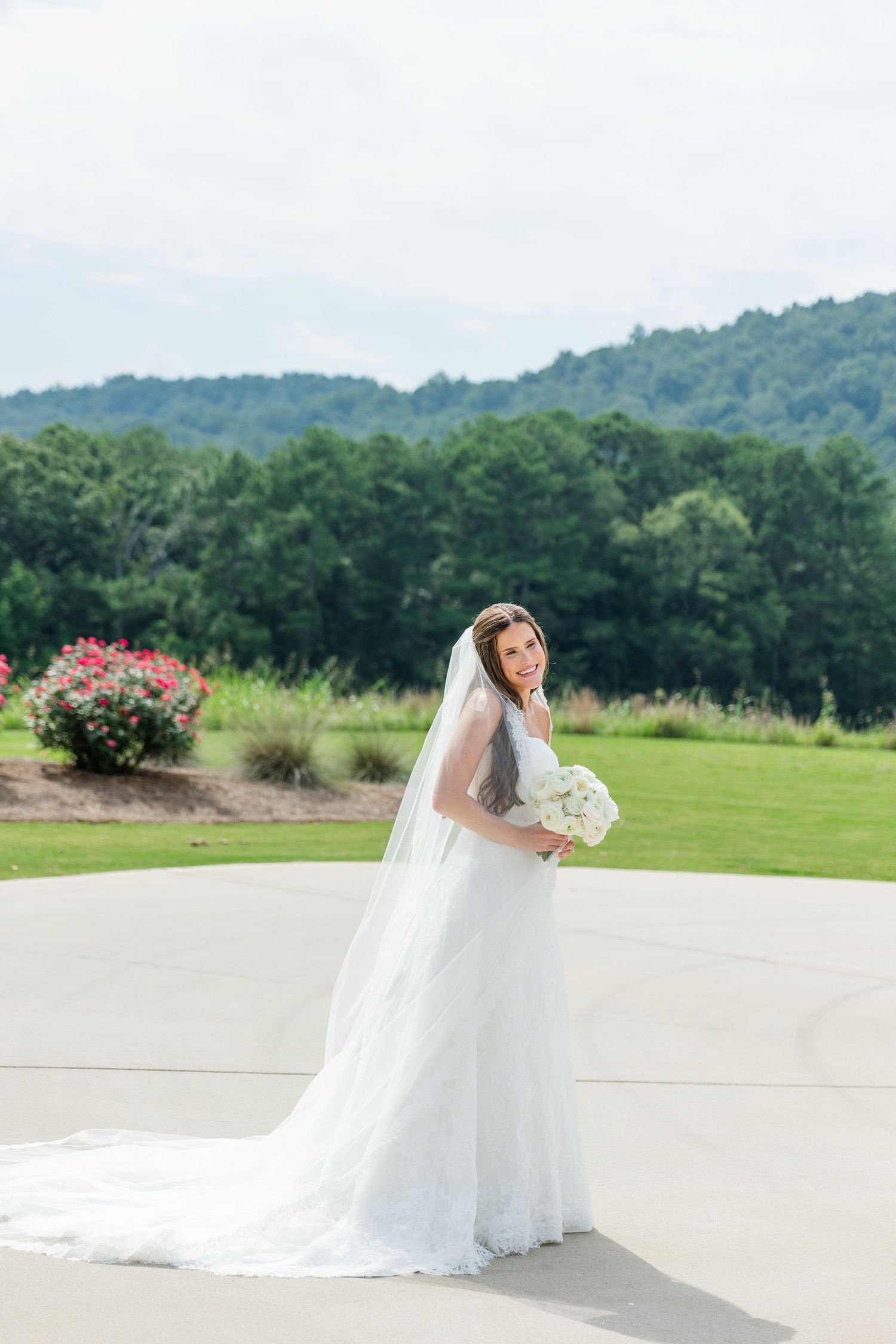 Bride in white gown and long veil holds bouquet, posing on patio with lush green hills and trees in background.