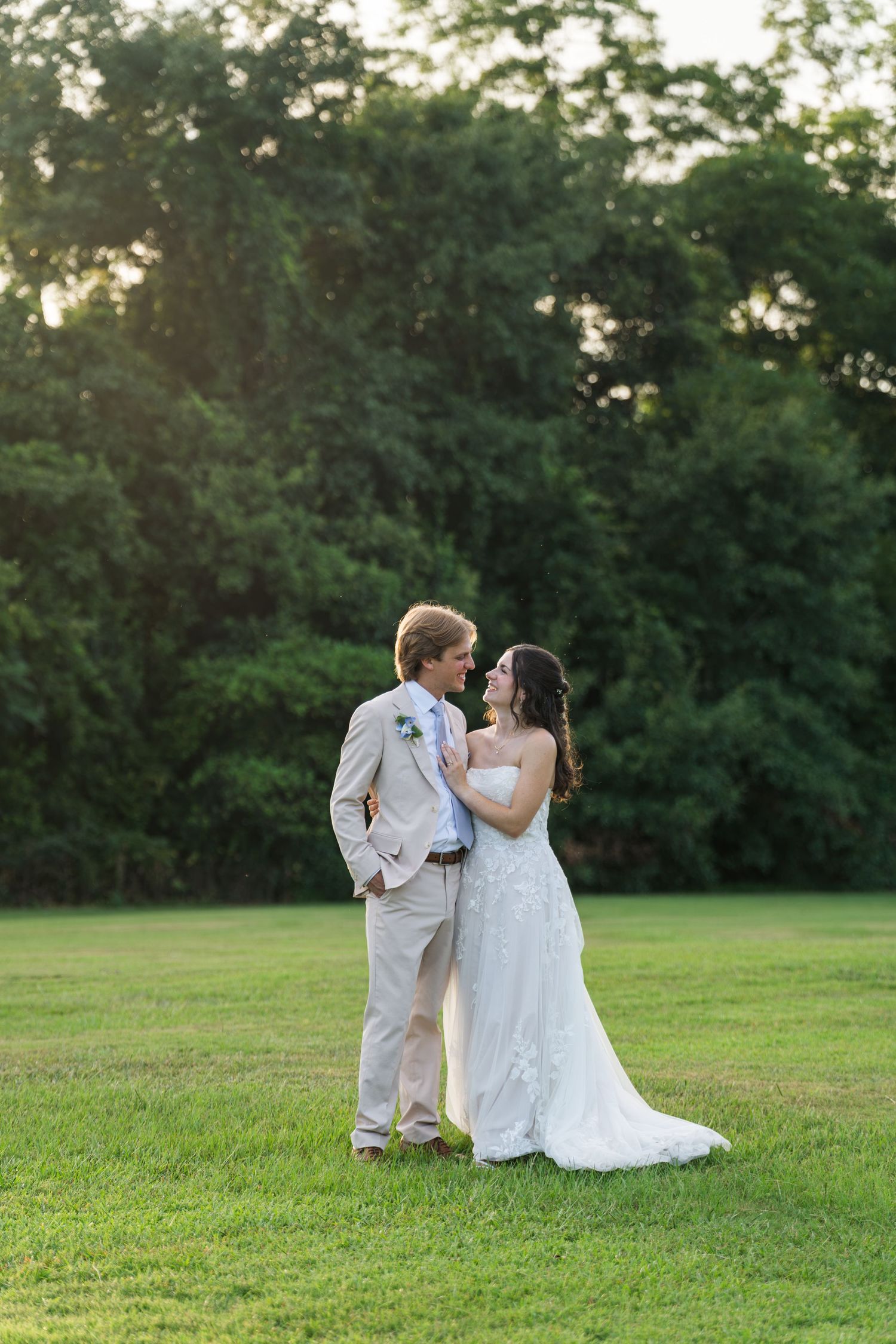 Bride and groom share a romantic moment on a lush green lawn at golden hour, surrounded by trees.