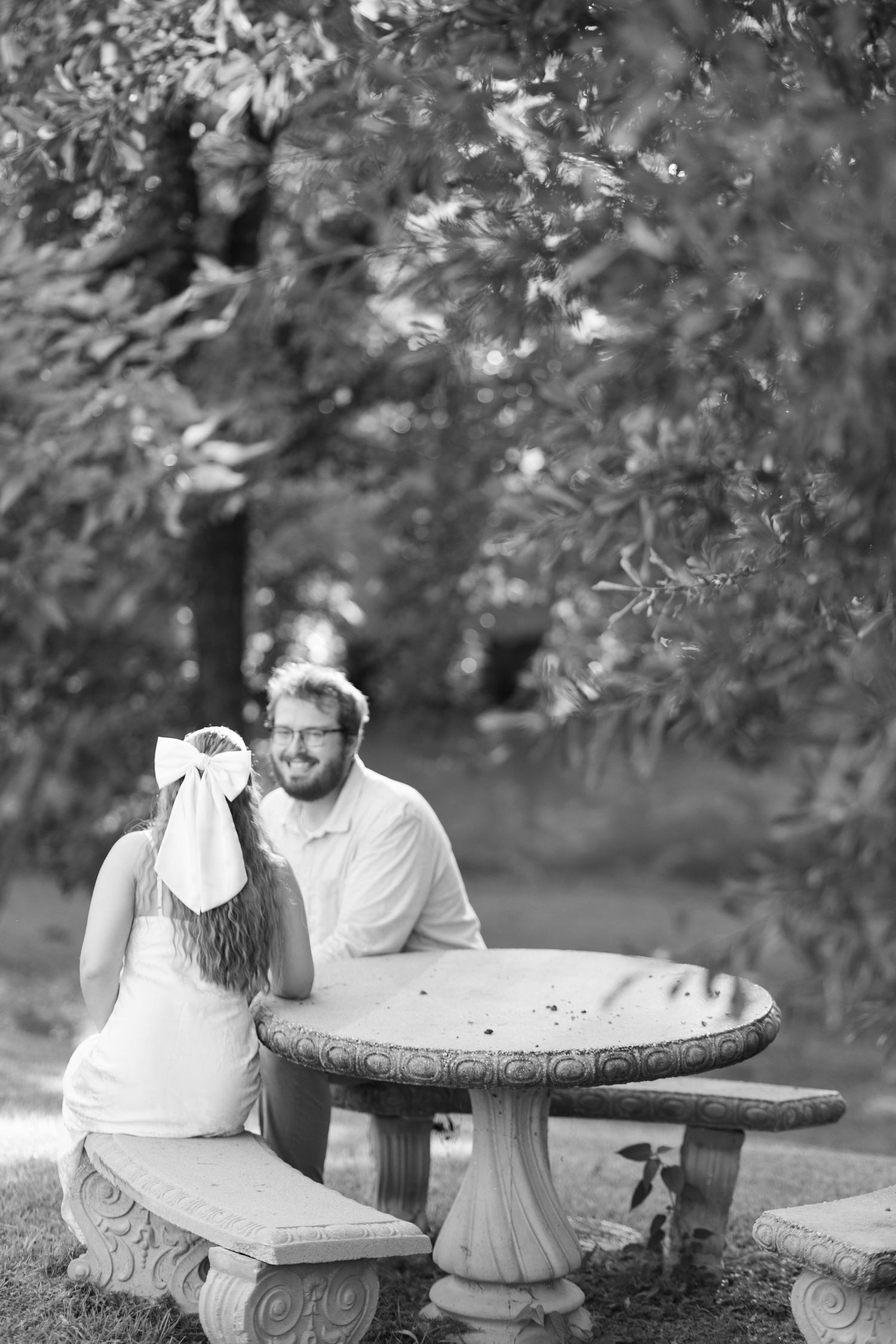 Couple sits at stone picnic table under trees in black and white wedding photo.