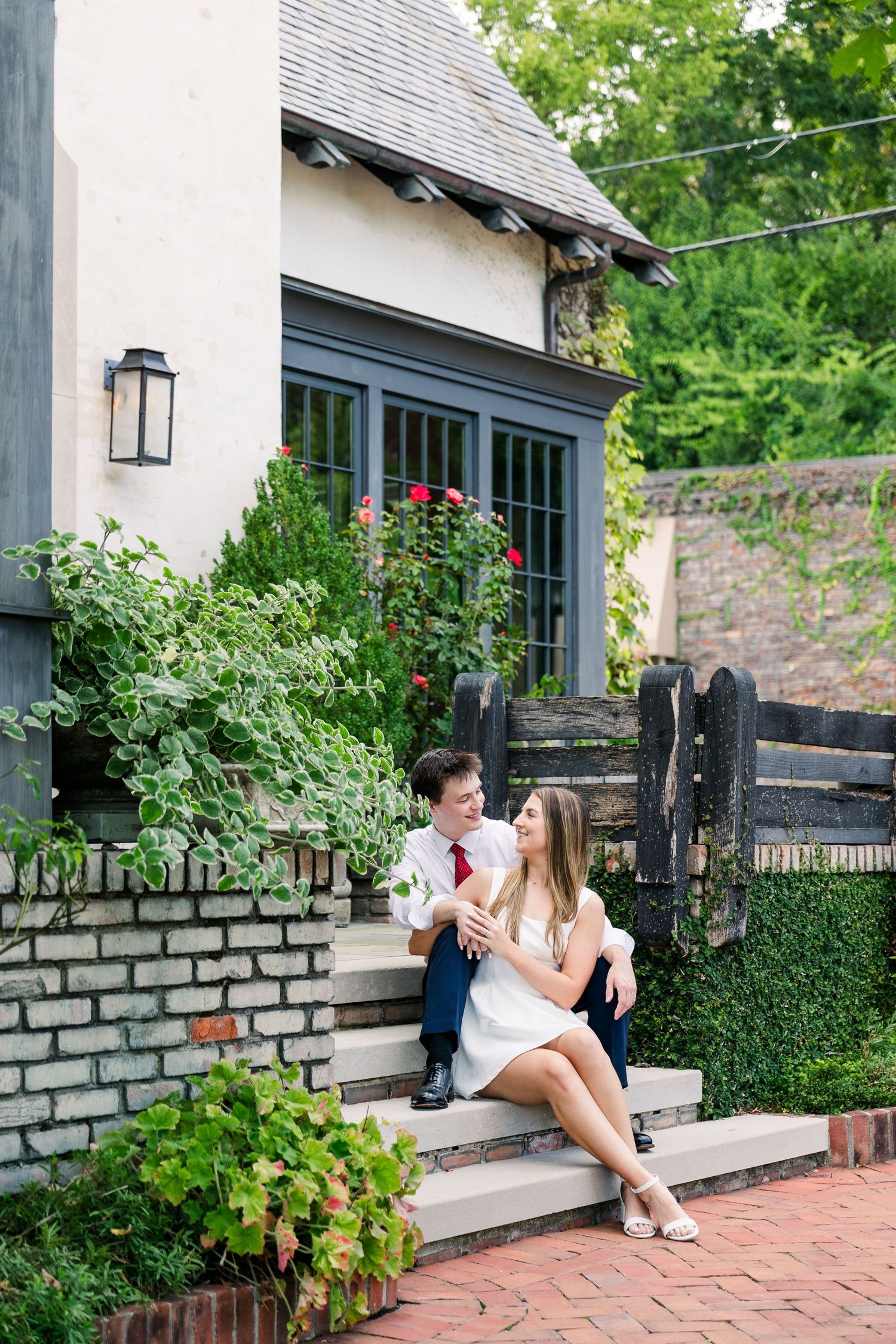 A couple sits on stone steps outside a charming Tudor-style home surrounded by lush greenery.