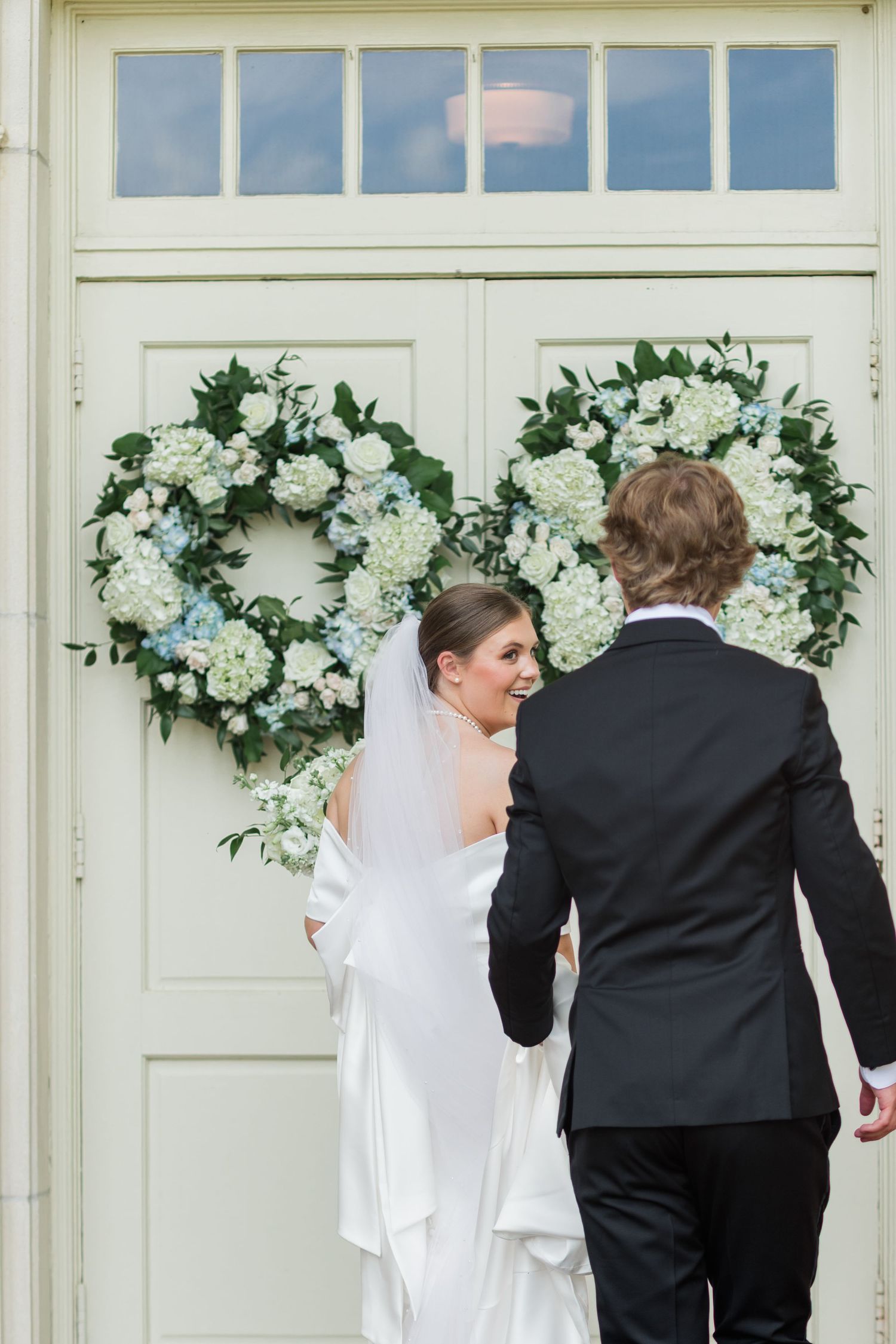 Bride in white gown and veil with groom entering church doors adorned with floral wreaths.
