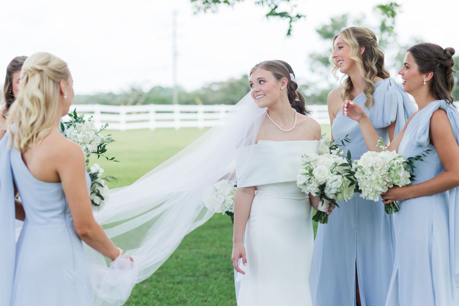 Bride in white gown with veil surrounded by bridesmaids in light blue dresses holding white floral bouquets outdoors.