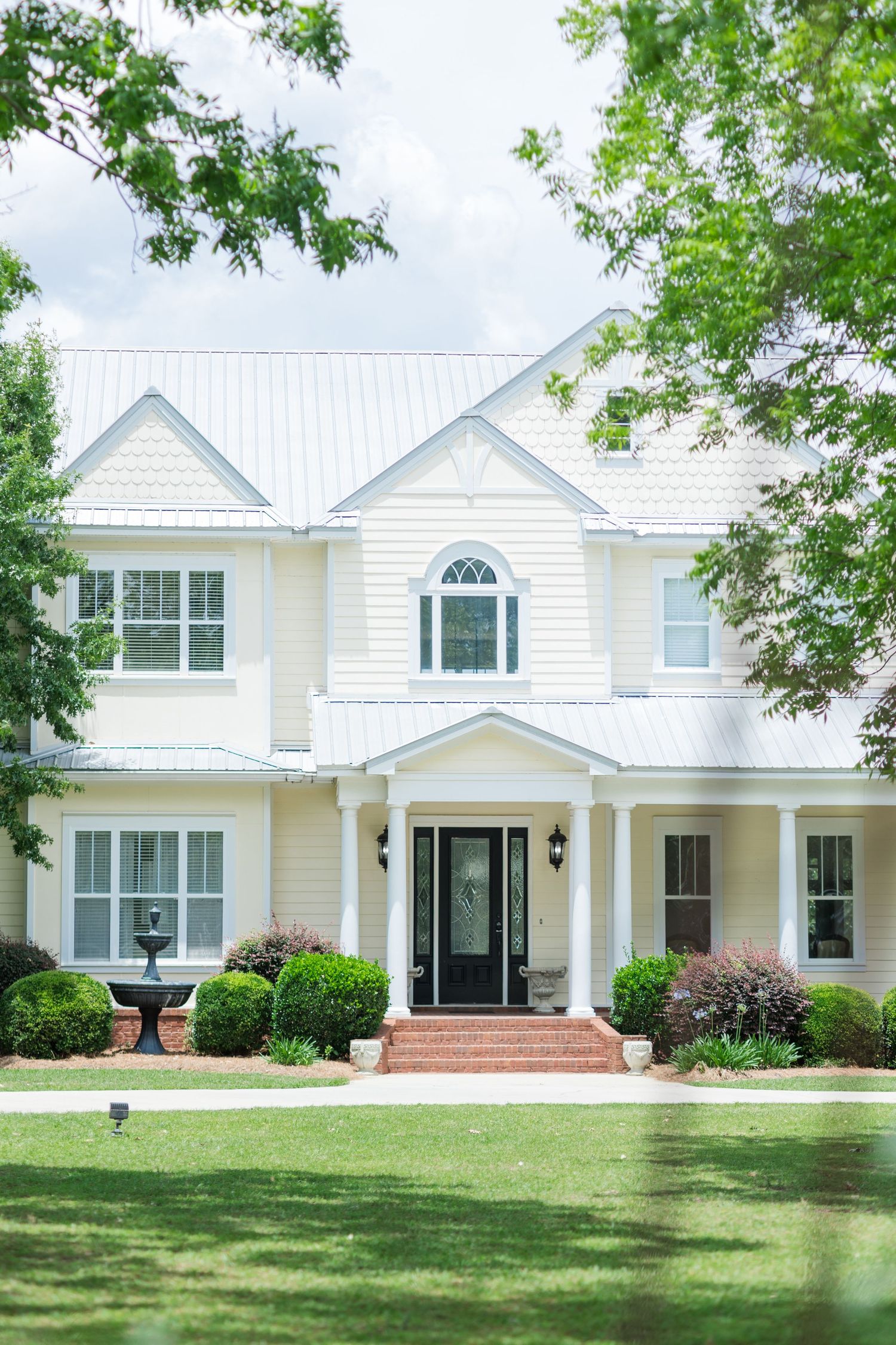 Elegant two-story white farmhouse with metal roof, brick steps, black front door, manicured shrubs, and lush green lawn.