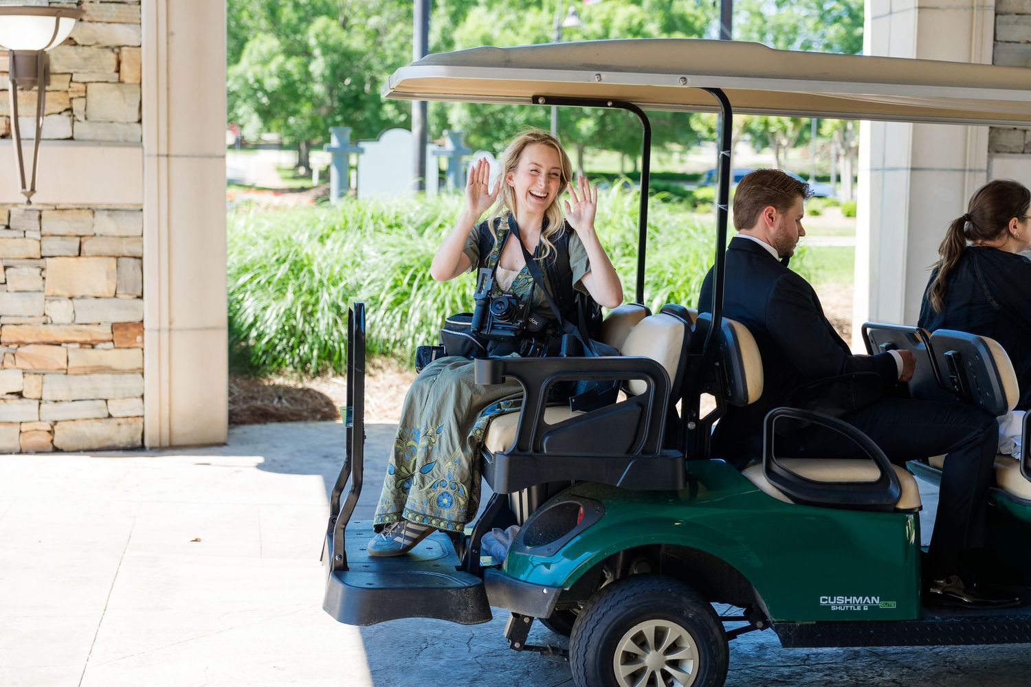 Woman in floral dress waves cheerfully while riding a green golf cart at an outdoor venue.