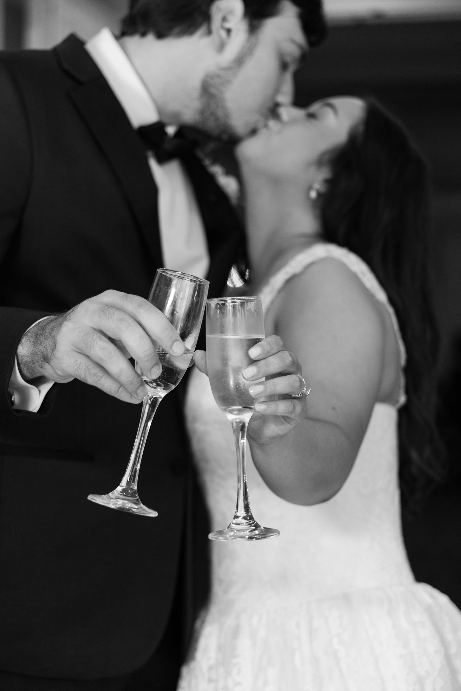 Black and white photo of newlyweds kissing while holding champagne flutes at their wedding reception.