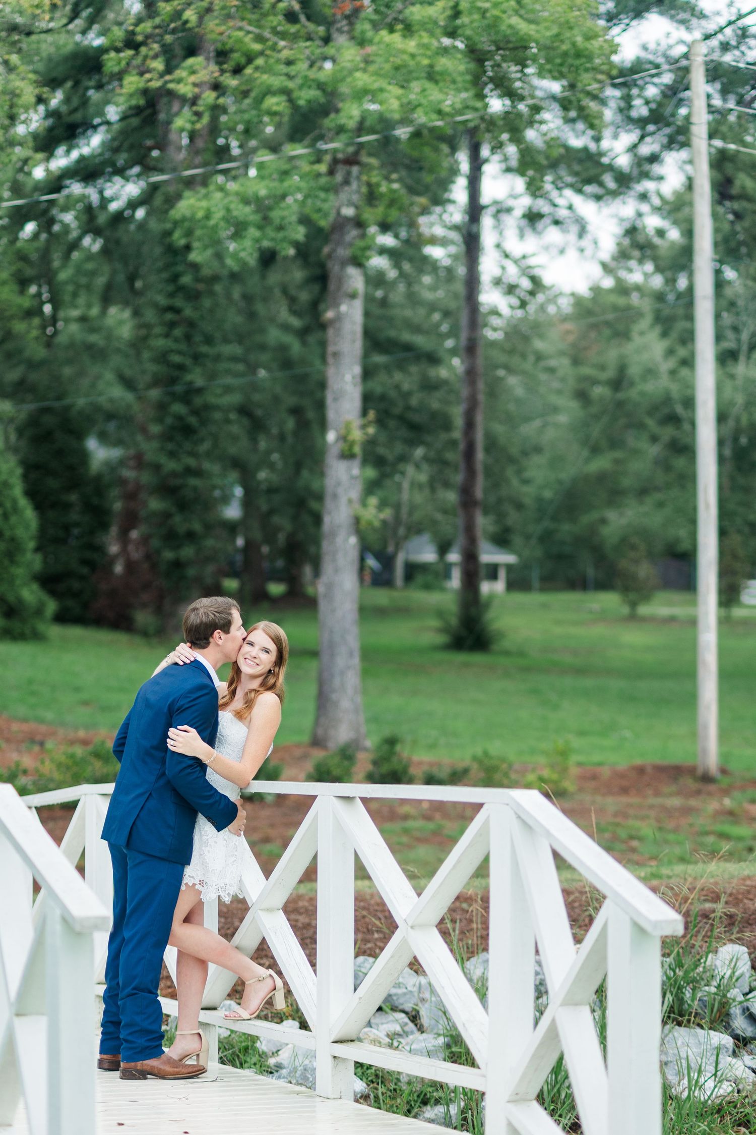 Couple sharing a romantic kiss on a white wooden bridge surrounded by lush green trees in an outdoor park setting.