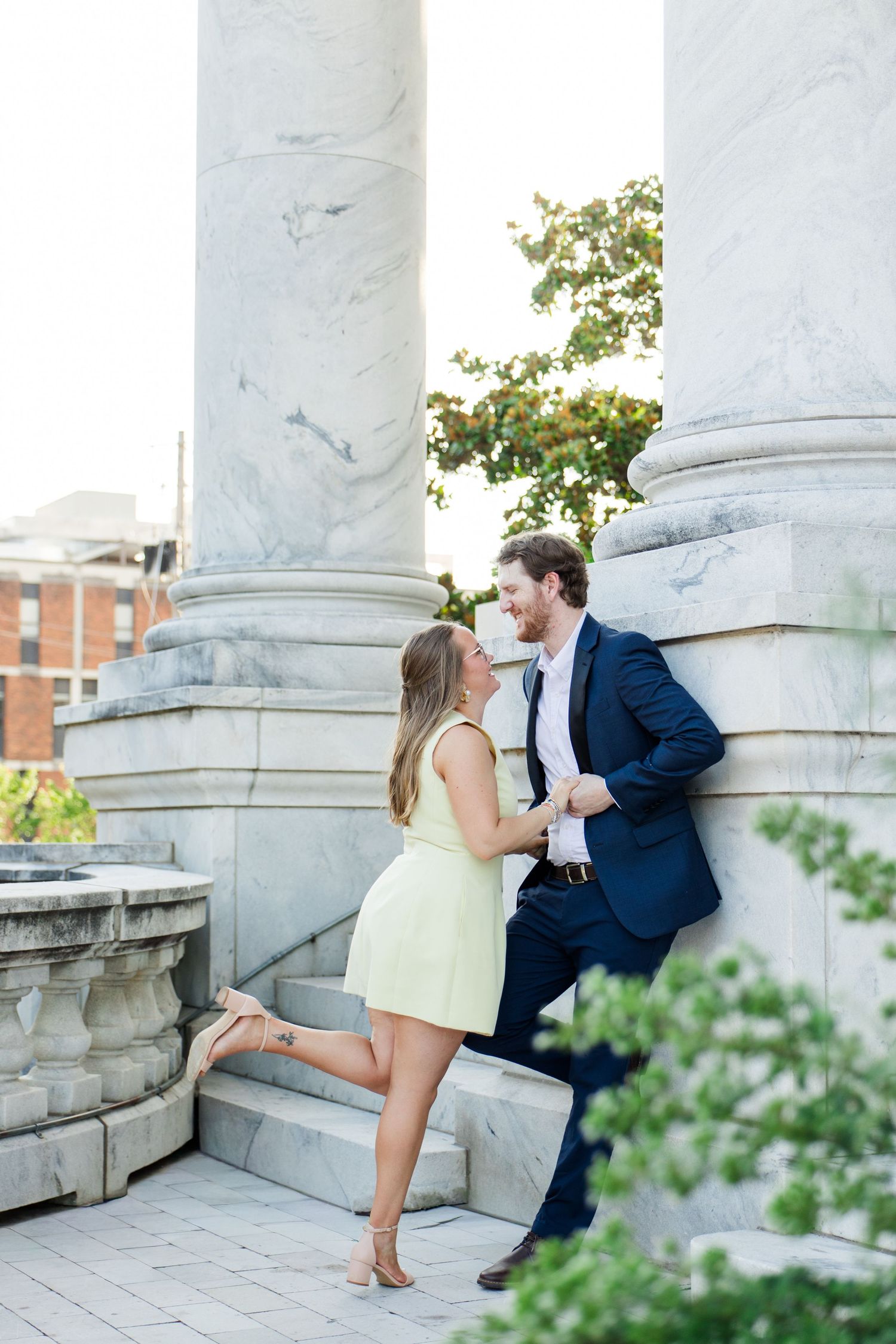 Couple laughing together by marble columns, woman in green dress and man in navy suit on stone steps outdoors.