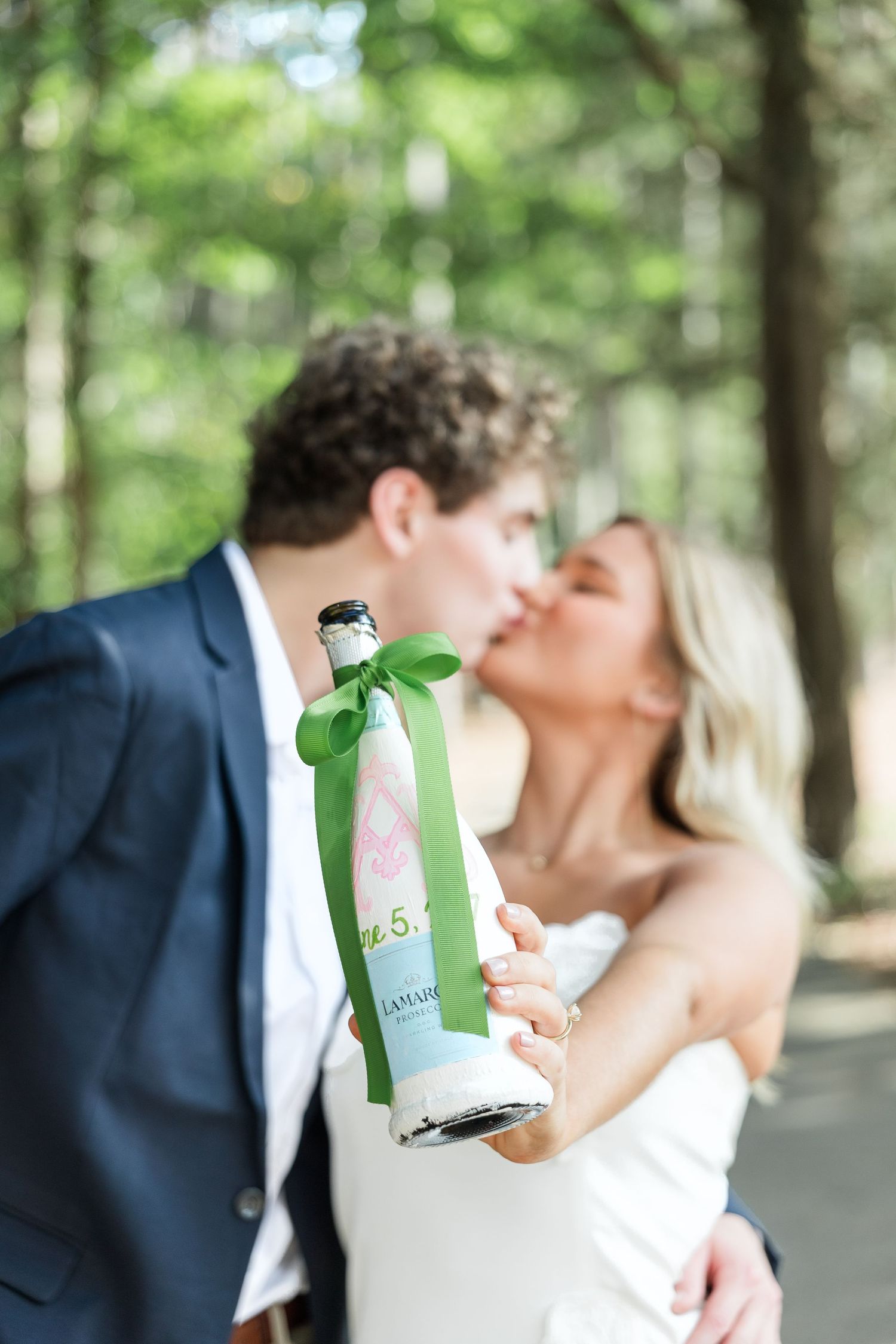 Wedding couple kisses while holding green ribbon-tied bottle in blurred forest background.