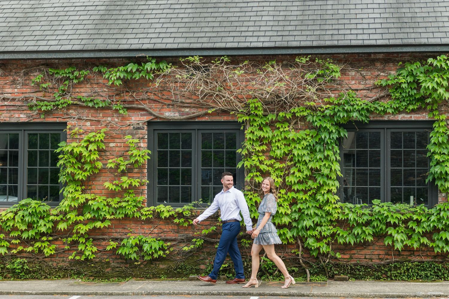 Couple walking hand in hand past a charming brick building covered in green ivy vines.