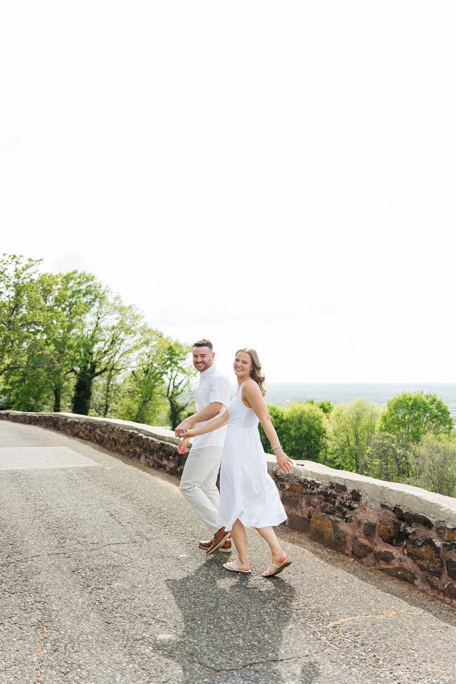 A couple in white outfits walk hand-in-hand along a scenic road with green trees and open sky.