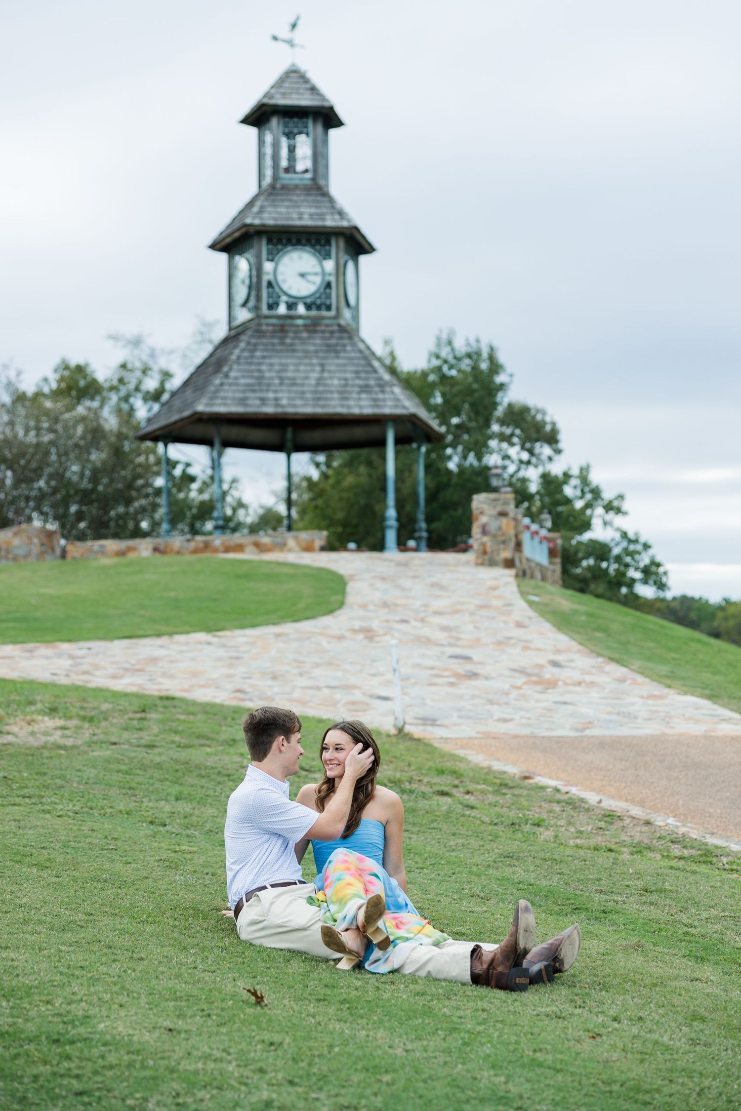 Couple sitting close together on a grassy hillside, smiling, with a stone pathway and a clock tower gazebo behind them.