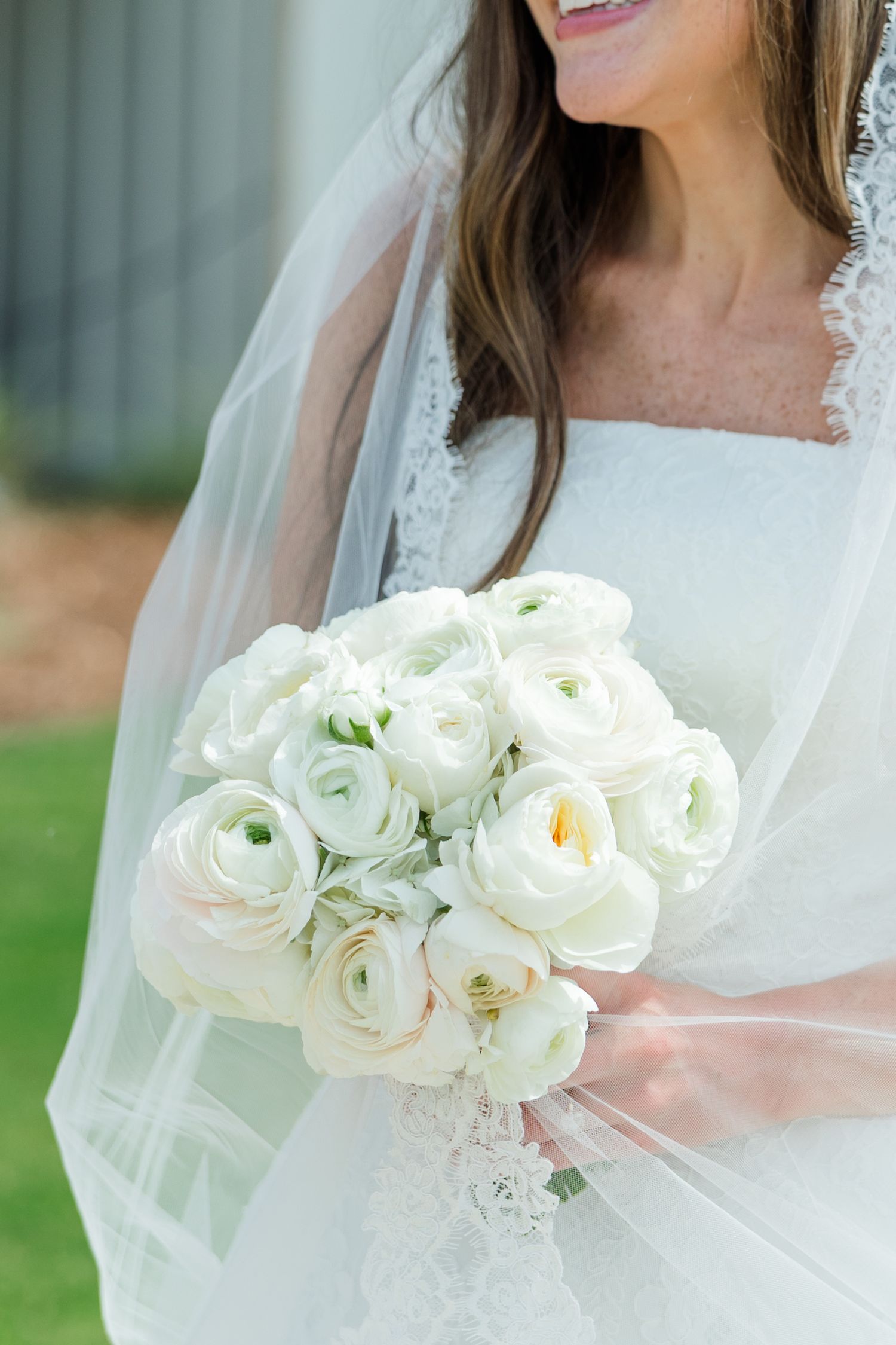 Bride holding a lush white ranunculus bouquet, wearing a lace wedding dress and veil.