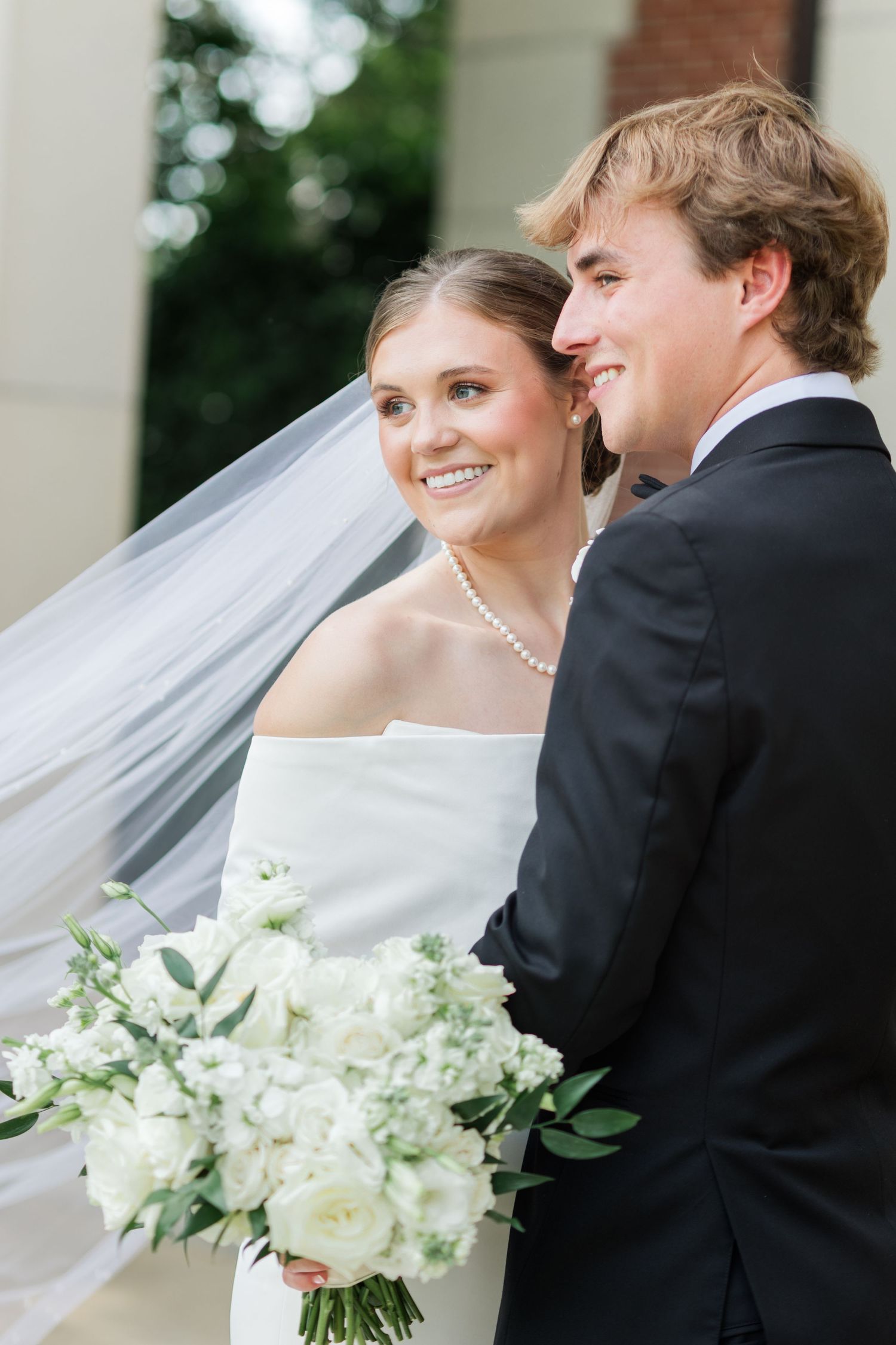 Elegant bride in off-shoulder gown with flowing veil holds white bouquet beside groom in black tuxedo.