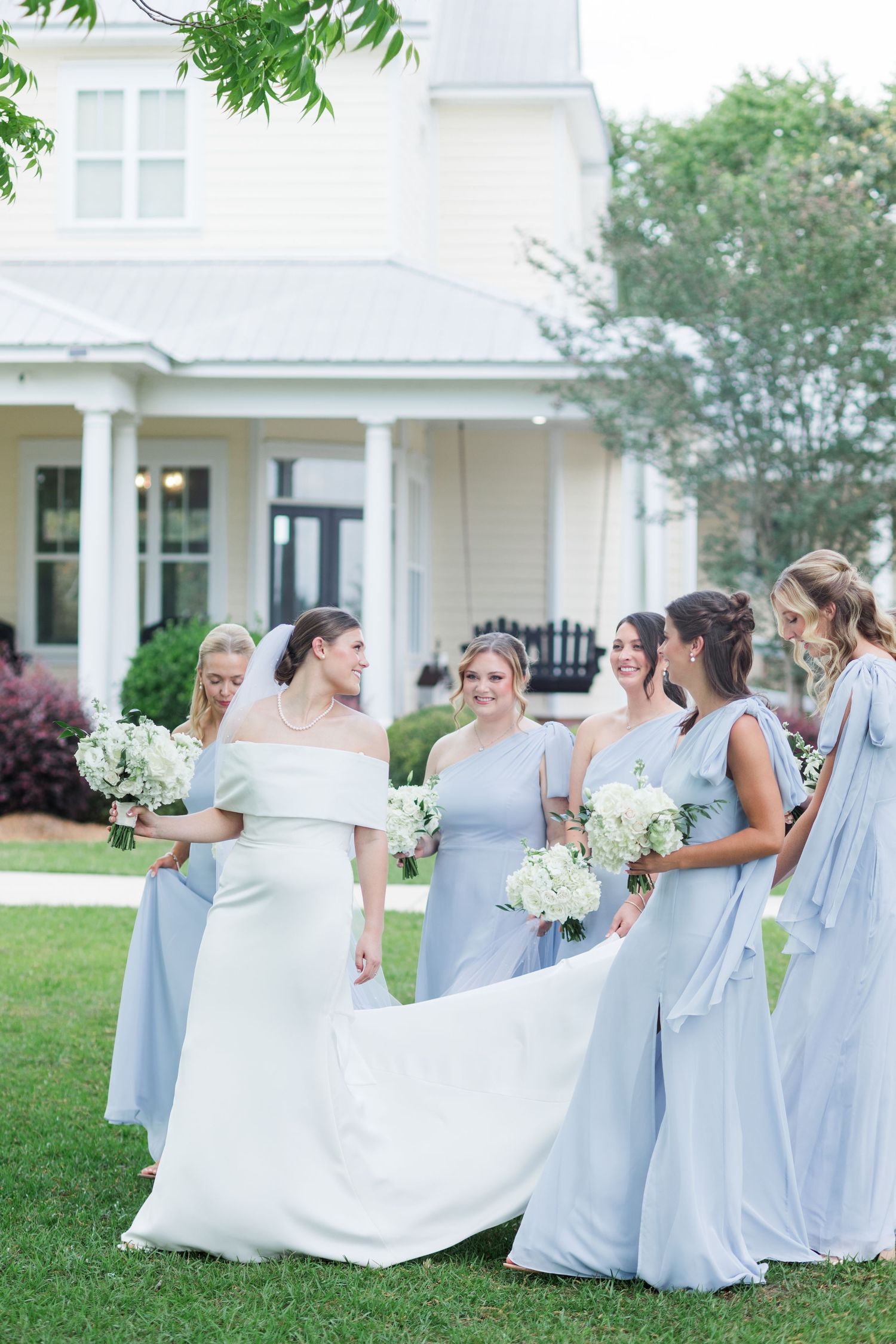 Bride in white gown surrounded by bridesmaids in light blue dresses holding white floral bouquets outside a white home.