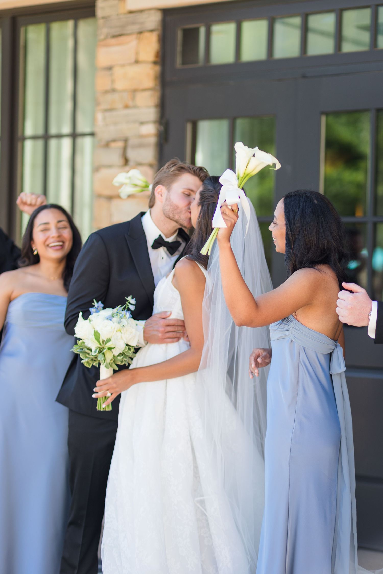 Bride and groom share a kiss surrounded by bridesmaids in blue gowns holding white floral bouquets outside a stone building.