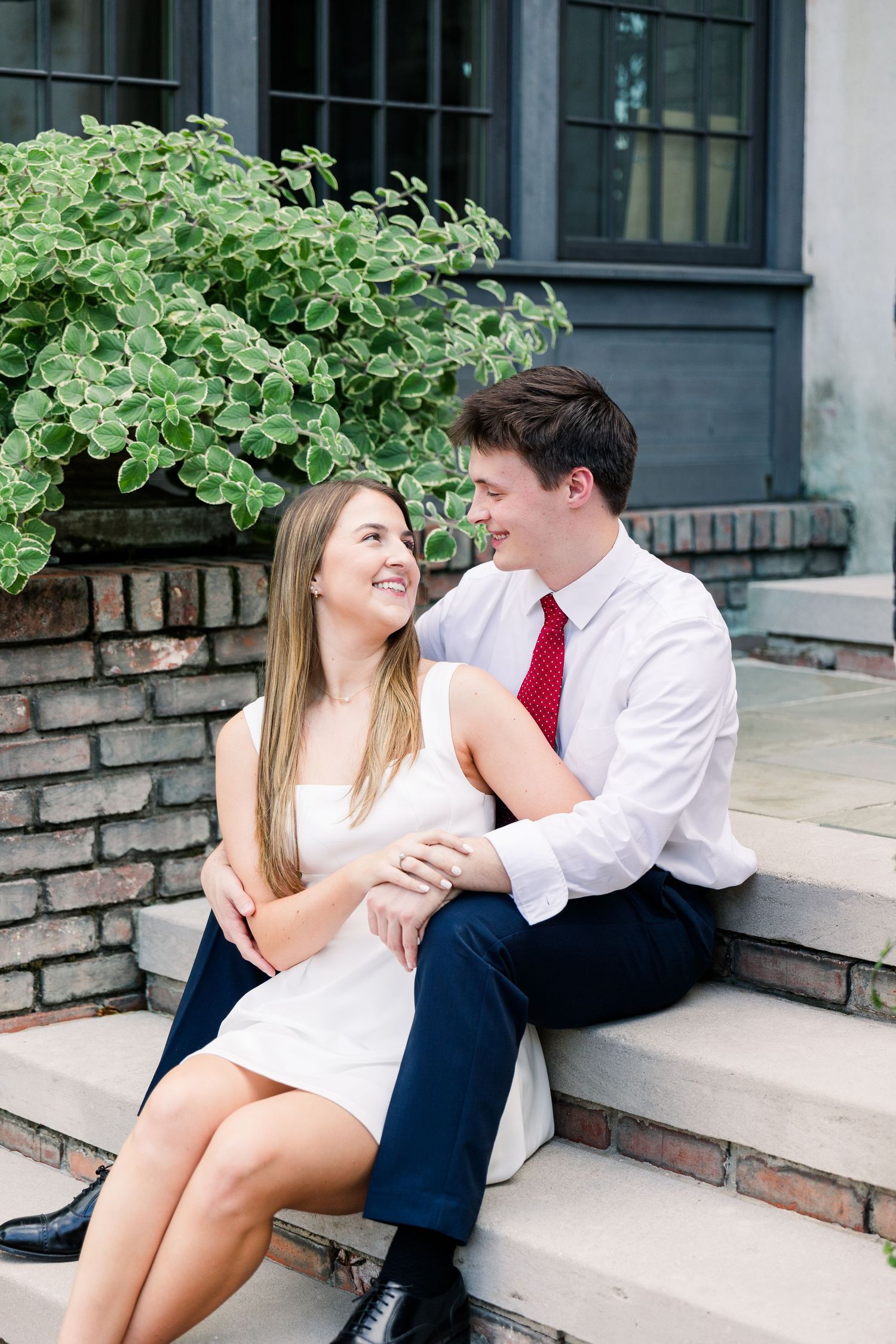 Engaged couple sitting on stone steps laughing together, woman in white dress, man in white shirt with red tie.