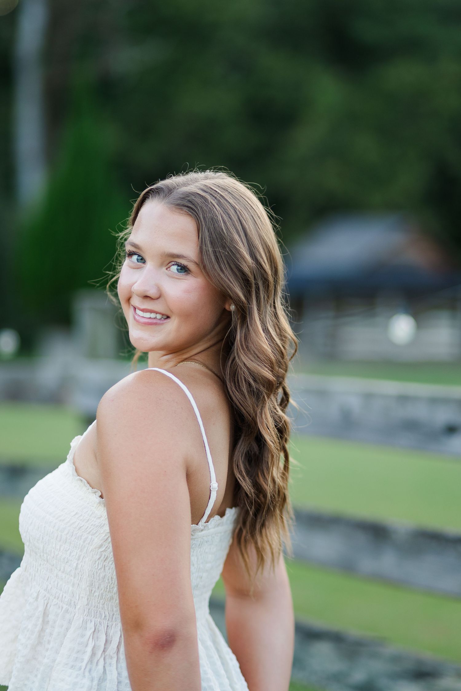 Smiling young woman in a white sundress with long wavy hair posing outdoors near a fence.