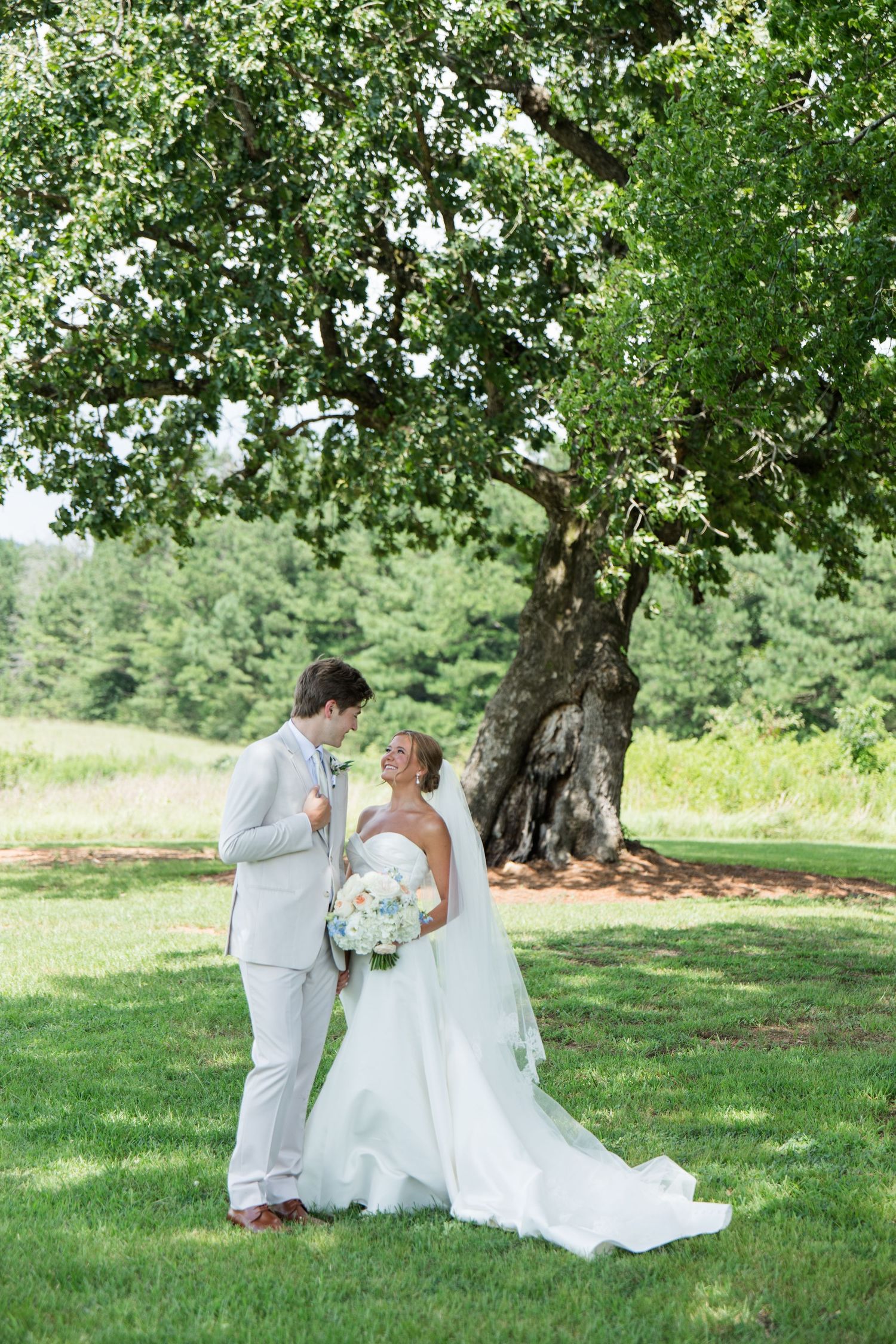 Bride and groom share a romantic moment under a large oak tree on a lush green lawn on their wedding day.