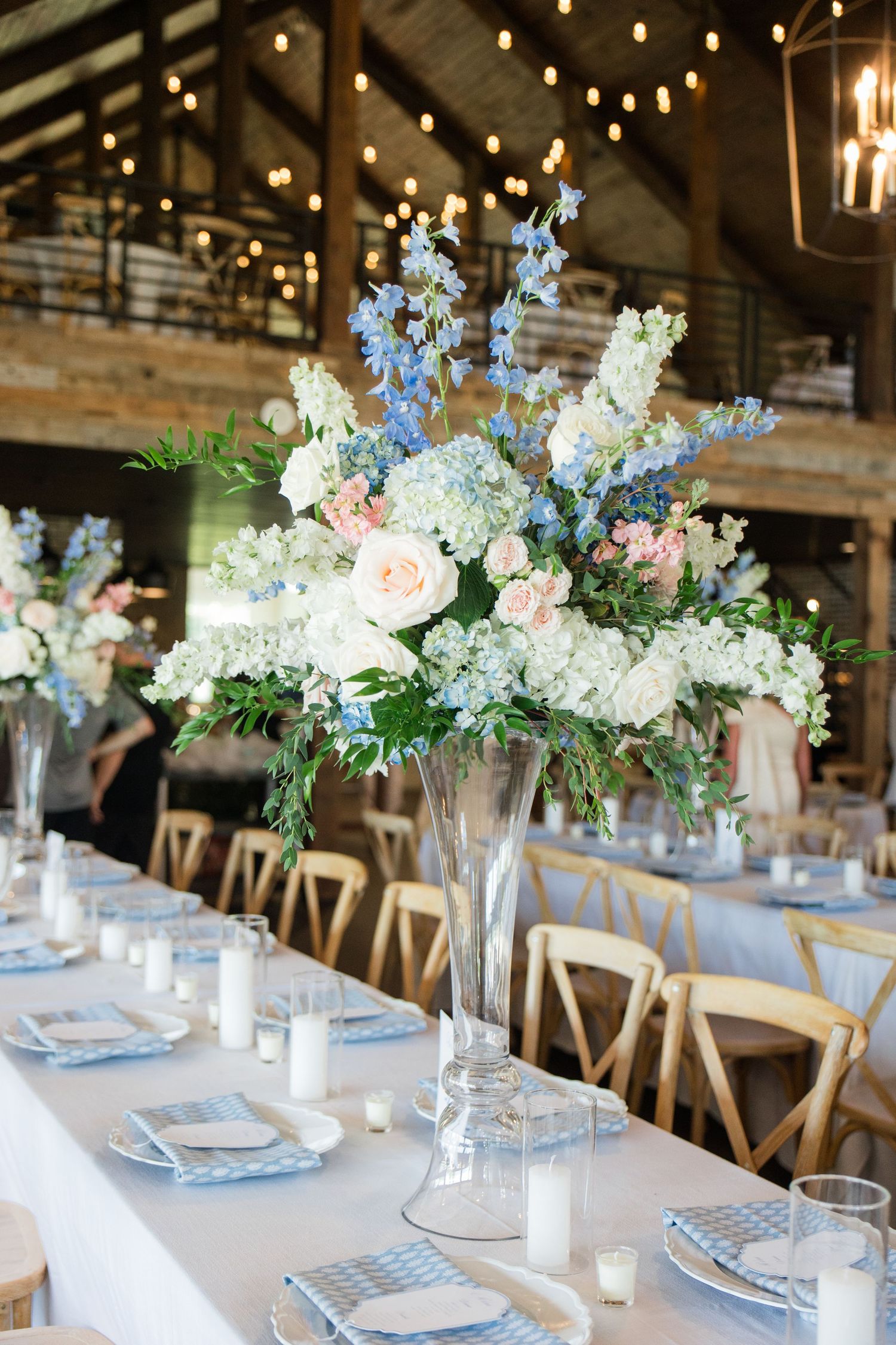 Elegant wedding reception table with tall floral centerpiece of blue, white, and blush flowers in a rustic barn venue.