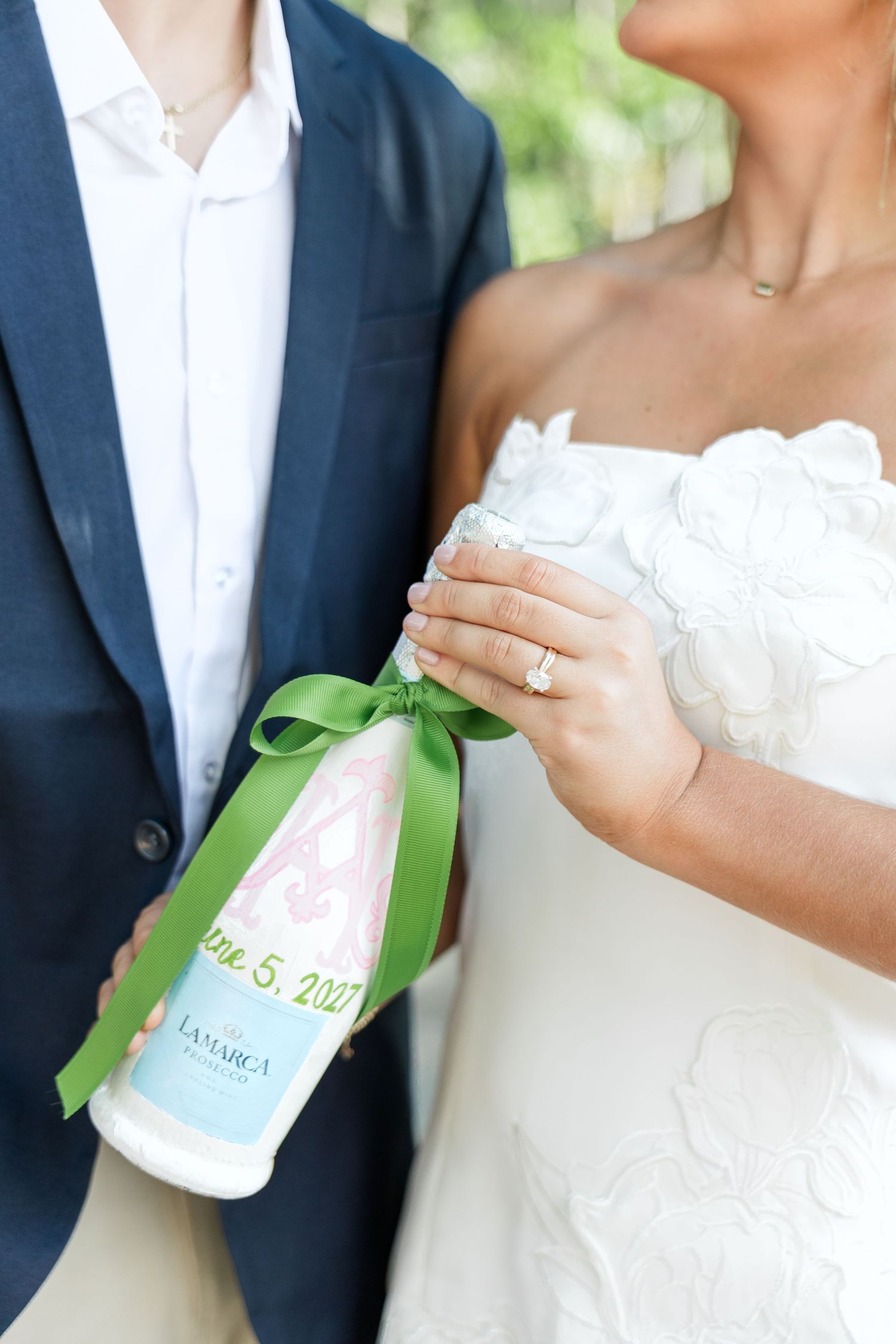 Close-up of bride and groom holding a La Marca champagne bottle with green ribbon and custom June 5, 2021 wedding label.