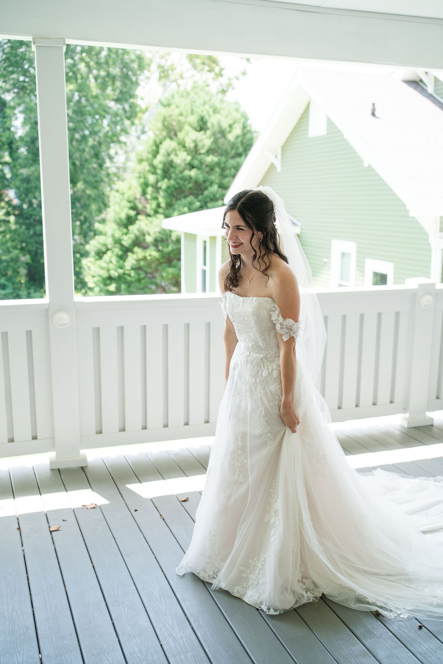 Bride in off-shoulder lace wedding gown with veil stands on white porch, smiling, with green building backdrop.