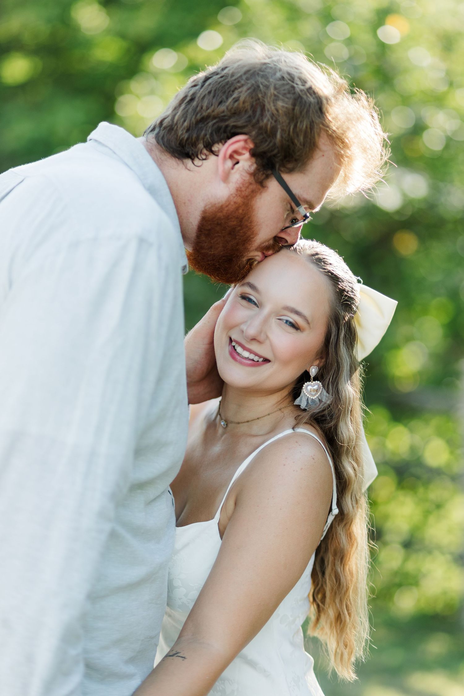 A smiling bride in white dress receives a kiss from a red-bearded groom outdoors in a lush green setting.