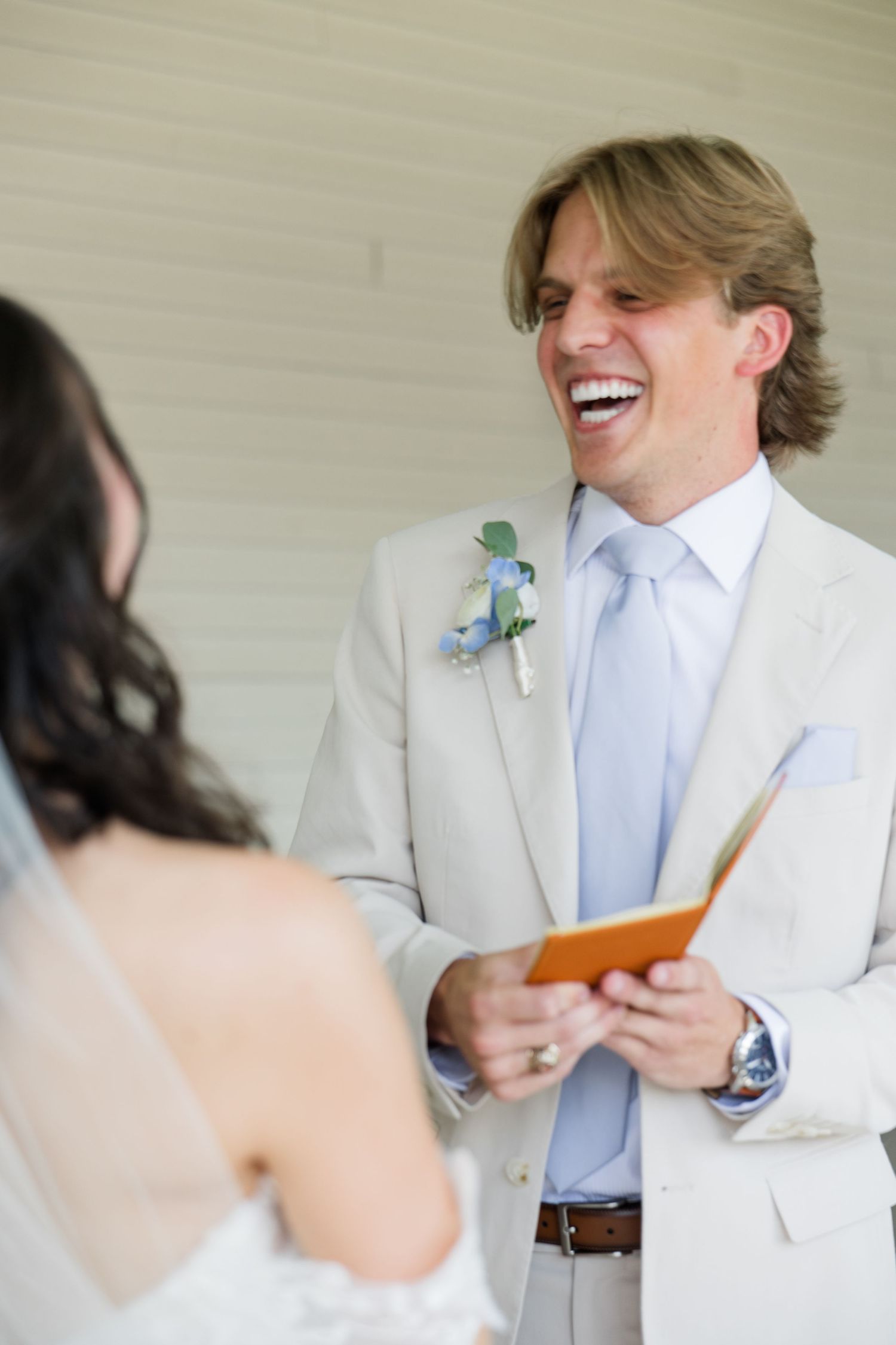 Joyful groom in tan suit laughs while reading wedding vows to his bride during their first look.