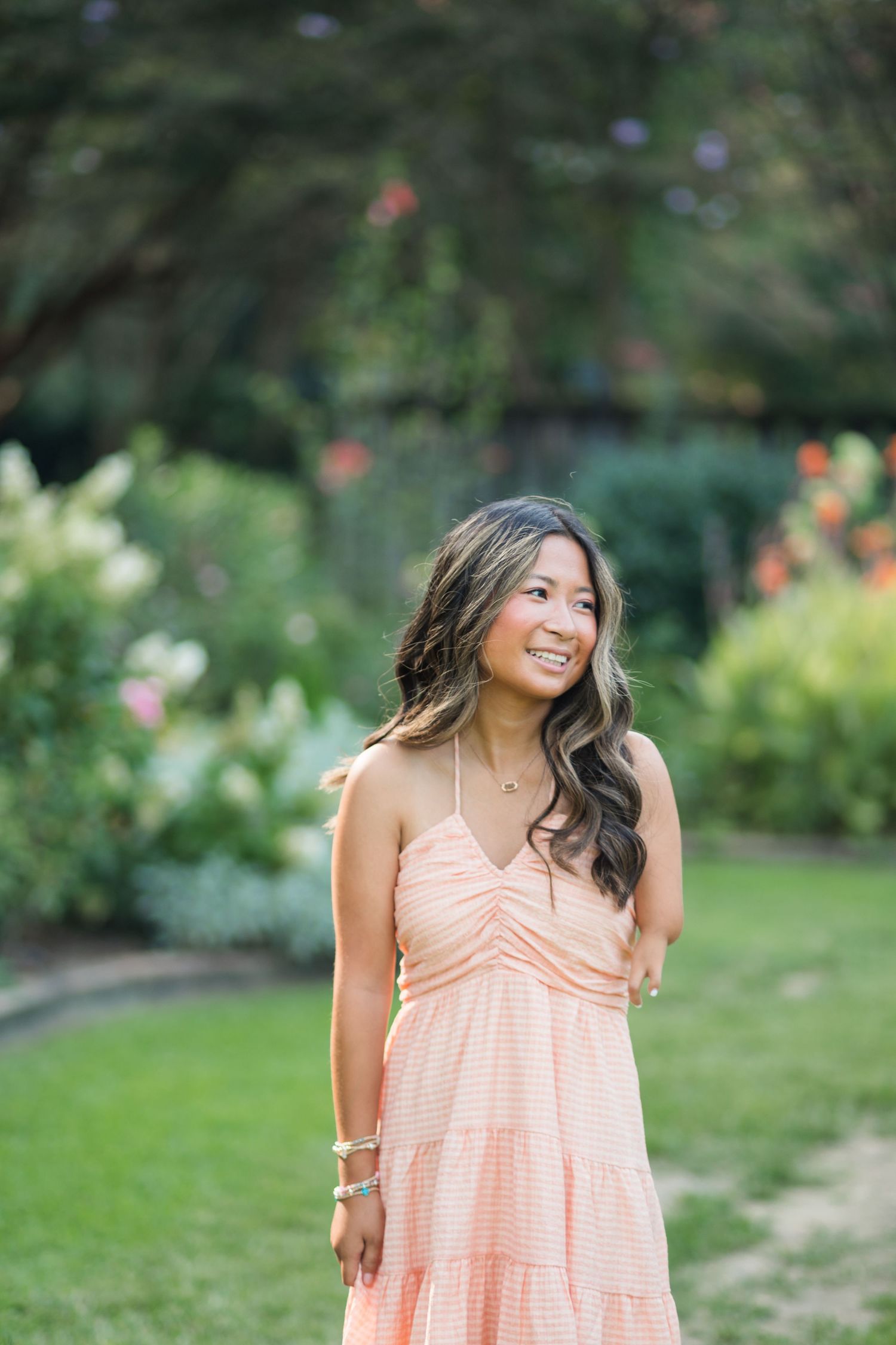 Young woman in peach sundress smiling in lush garden setting with colorful flowers and greenery.