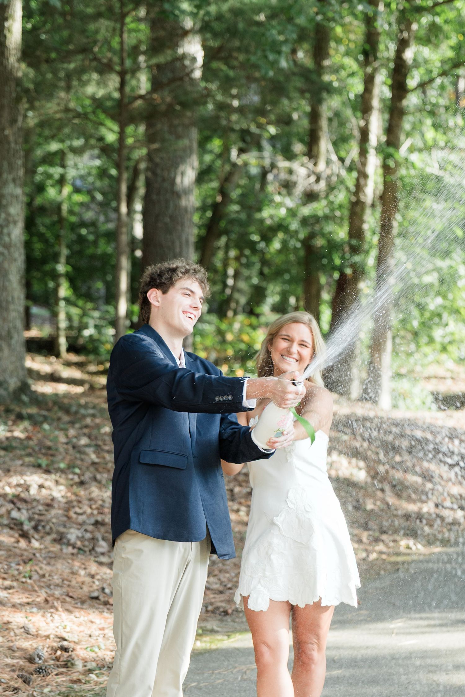 A joyful couple pops champagne outdoors, laughing as bubbles spray, dressed in a navy blazer and white dress.