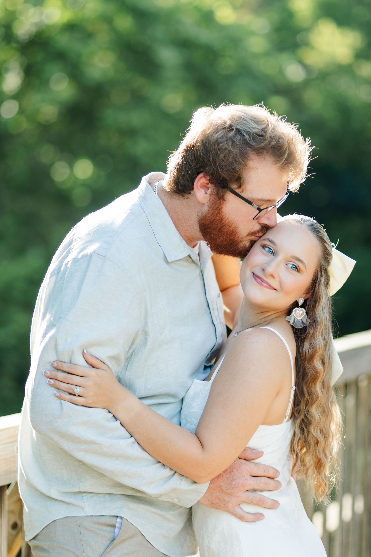 A bearded man in glasses kisses a smiling blonde woman in a white dress on a wooden deck outdoors.