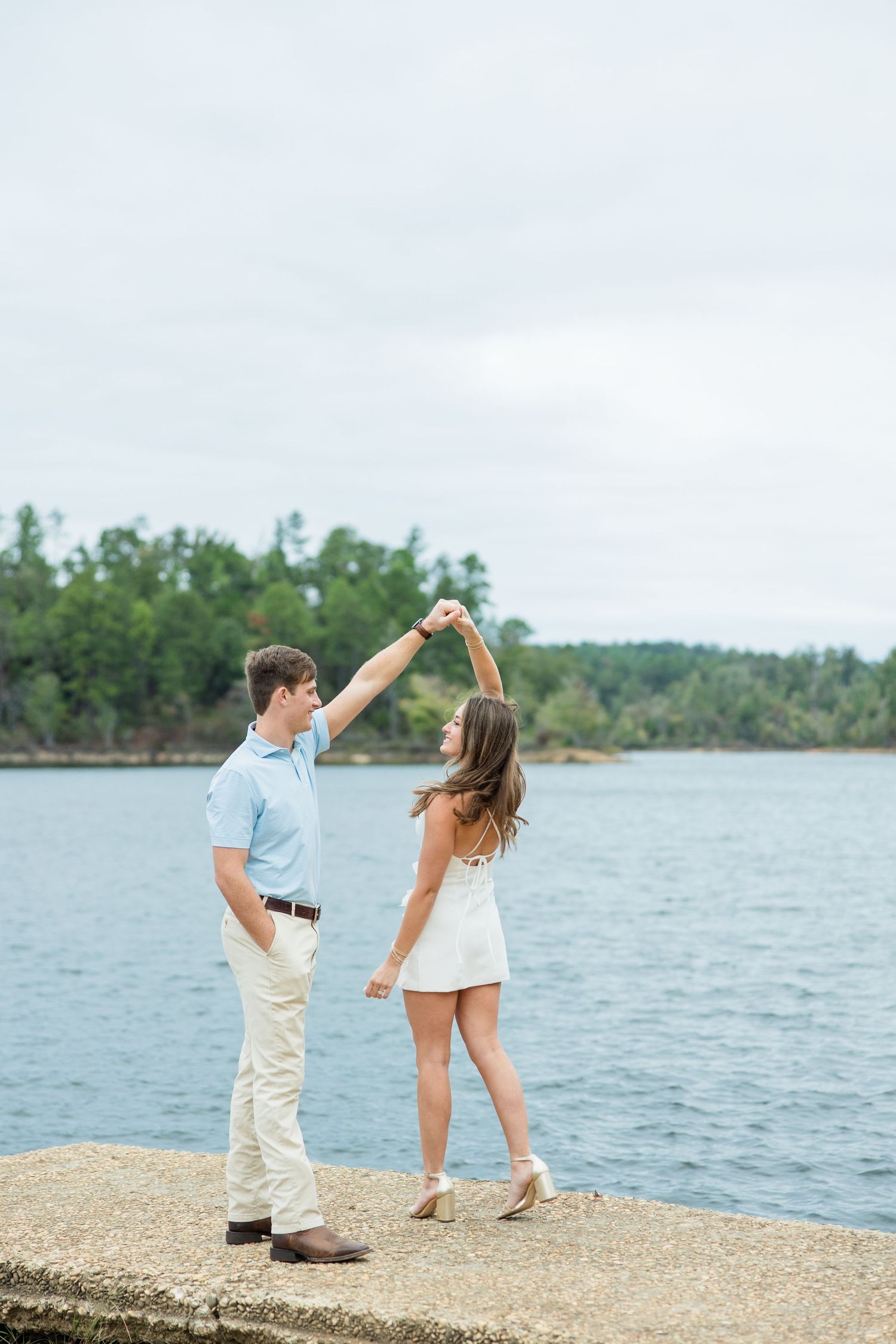 A man in a light blue shirt twirls a woman in a white dress on a stone lakeside pier with forested shores behind them.