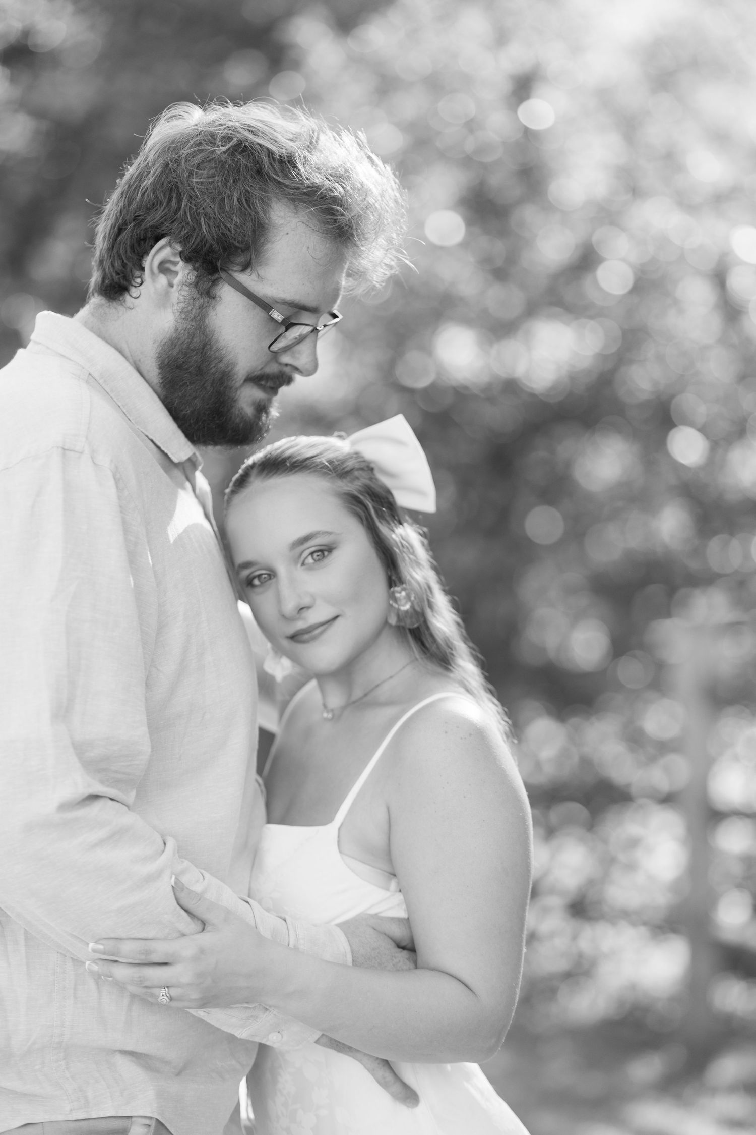 Black and white portrait of a couple embracing outdoors, woman in white dress with bow smiling softly at camera.