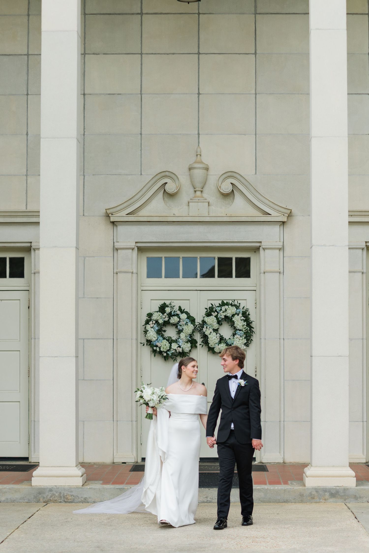 Bride and groom holding hands walking before a grand white columned building decorated with matching floral wreaths.