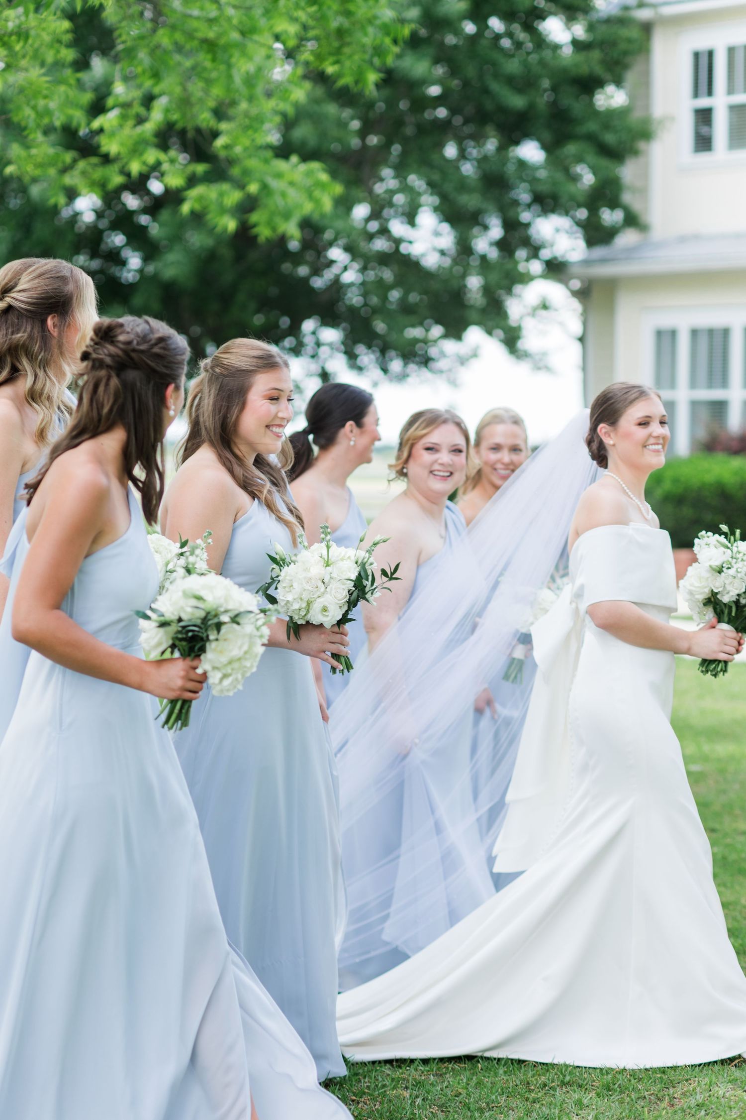 Bride in white off-shoulder gown walks with bridesmaids in light blue dresses outdoors.