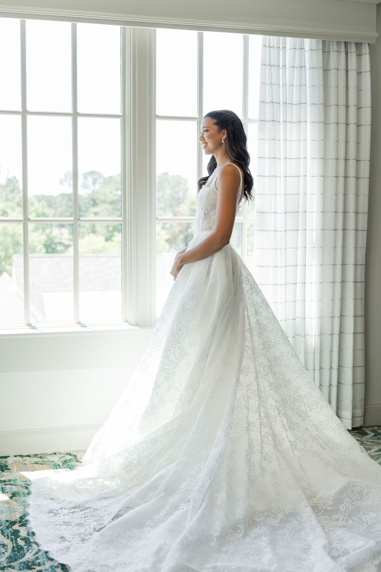 Bride in elegant lace wedding gown with long train stands by large window in soft natural light.