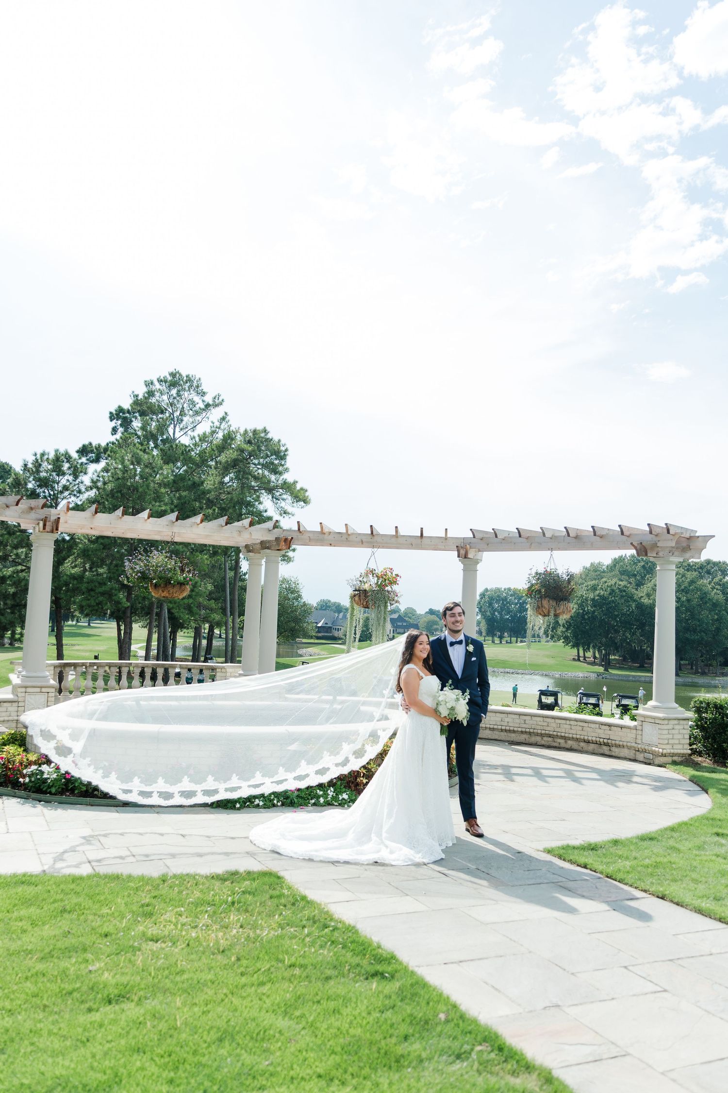 Bride and groom pose on stone pathway with flowing veil and pergola backdrop at outdoor wedding venue.