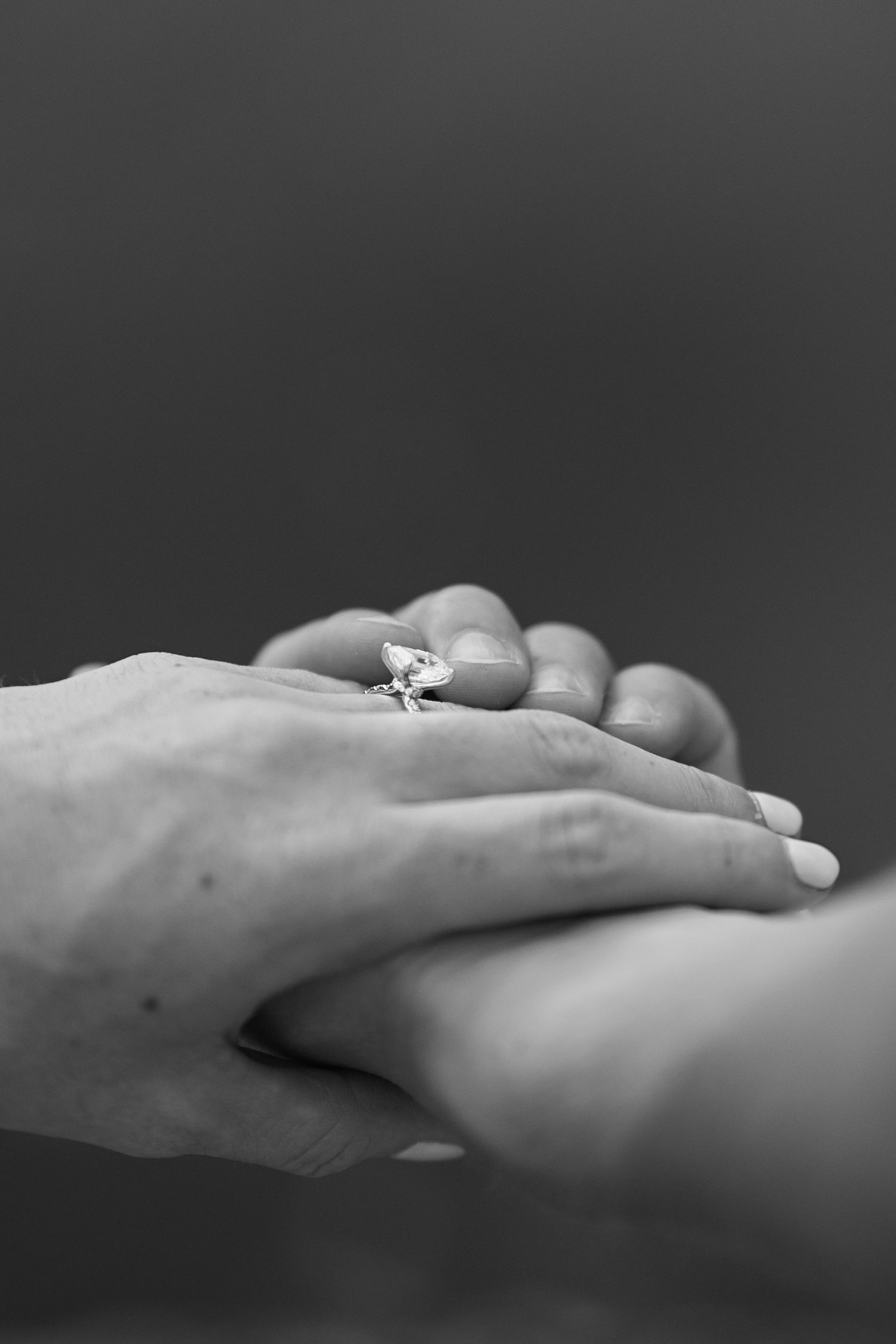 Black and white photo of two hands touching, one wearing a diamond engagement ring, symbolizing love and commitment.