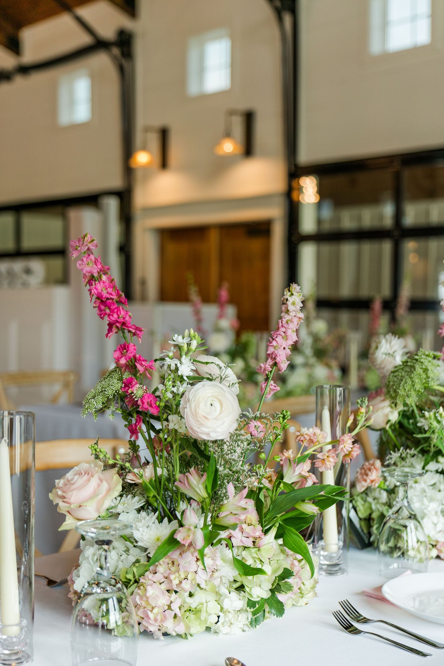 Elegant wedding reception centerpiece with pink and white flowers, greenery, and candles on a white tablecloth in a barn venue.