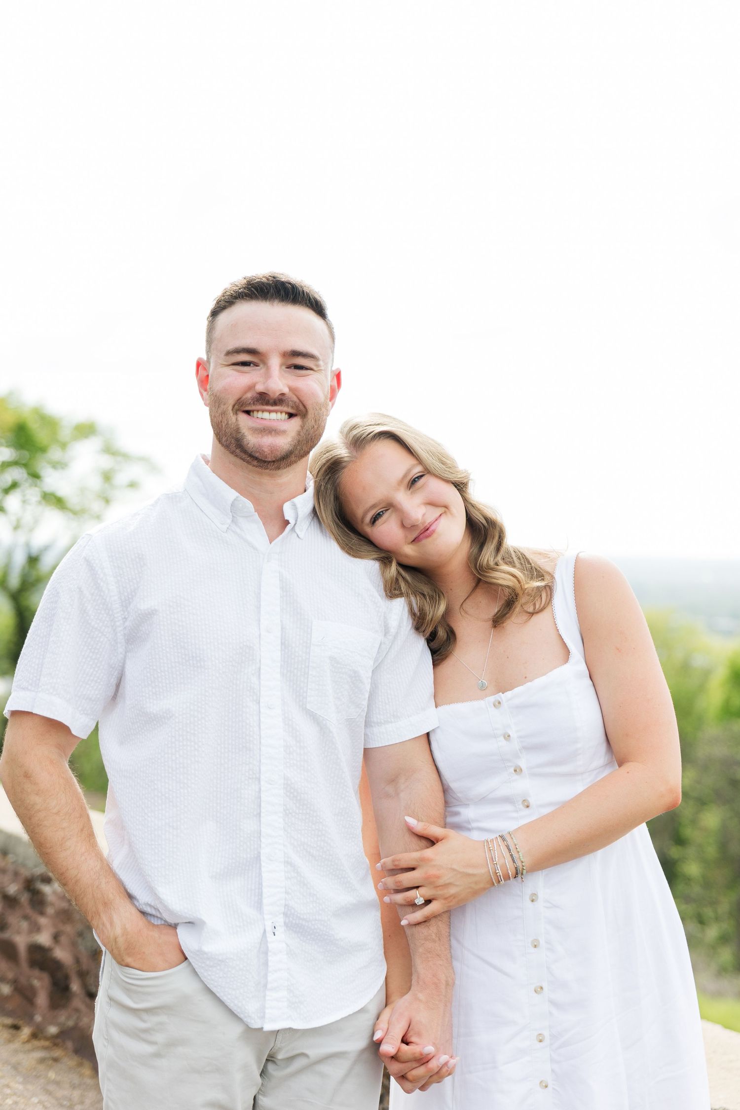 Smiling couple in white outfits holding hands outdoors with greenery in background.