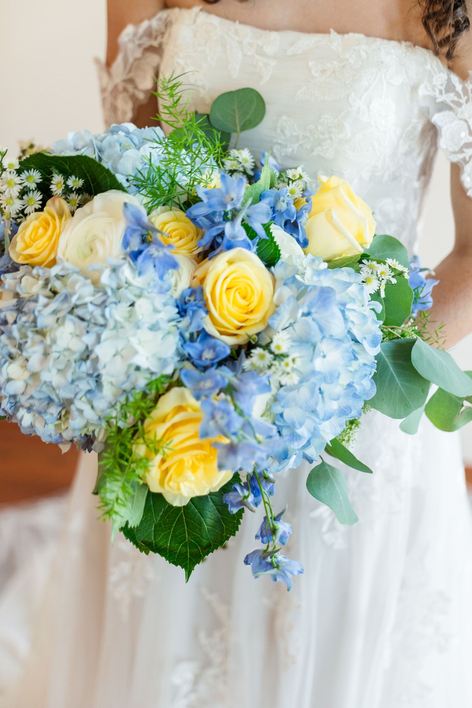 Bride holding cascading wedding bouquet with blue hydrangeas, yellow roses, and eucalyptus greenery.