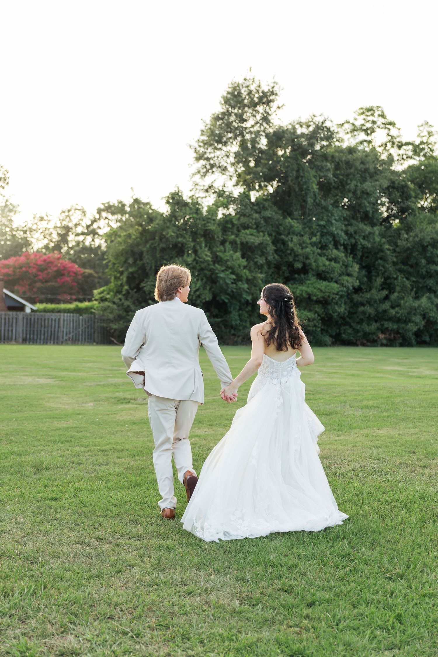 Newlywed couple holding hands walking across lush green lawn at sunset in wedding attire.