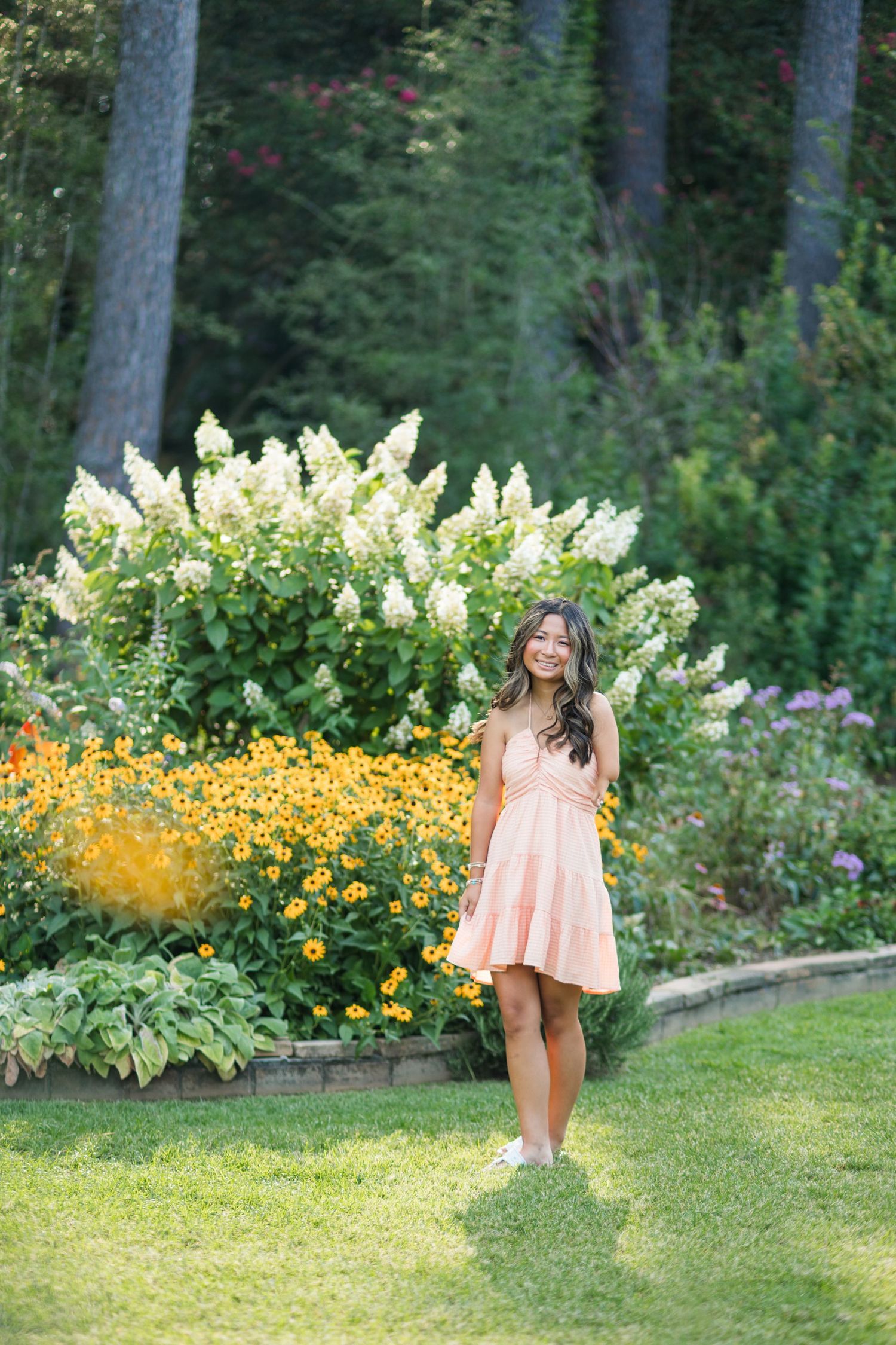 Young woman in a pink dress smiling in a lush garden with yellow flowers and white hydrangeas.