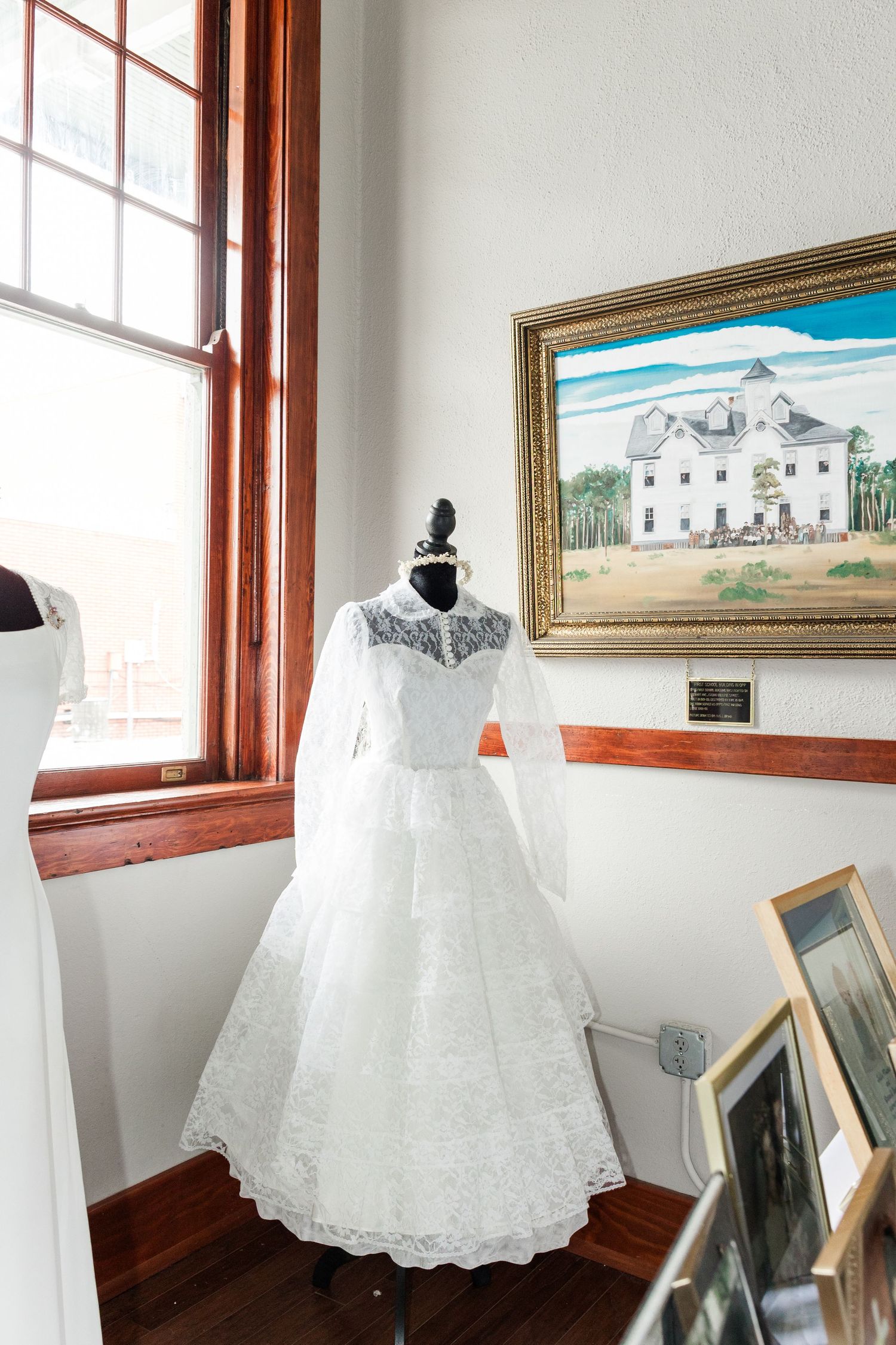 Vintage white lace wedding dress displayed on mannequin near window with framed painting of historic building on wall.