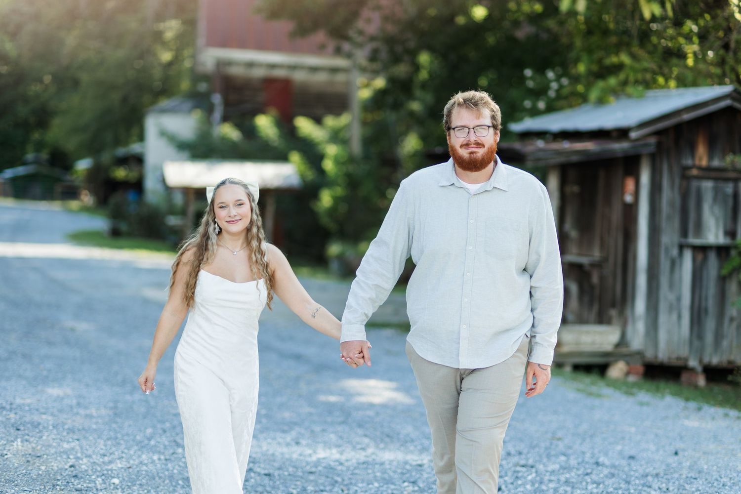 Couple holding hands walking on gravel path, woman in white dress, man in light blue shirt, rustic buildings in background.